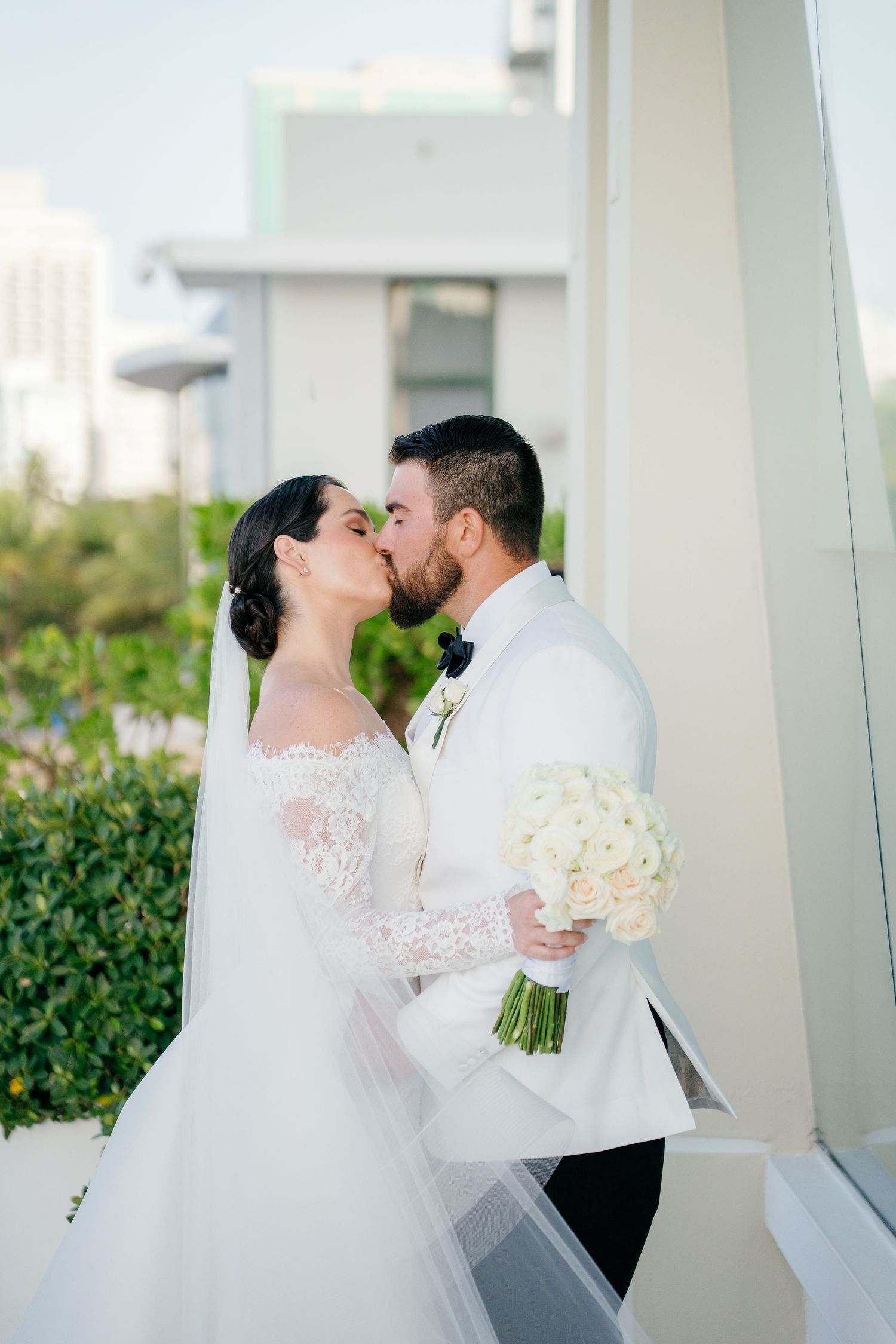 Bride and groom share a romantic kiss outdoors, bride holding white rose bouquet in elegant lace wedding gown with veil.