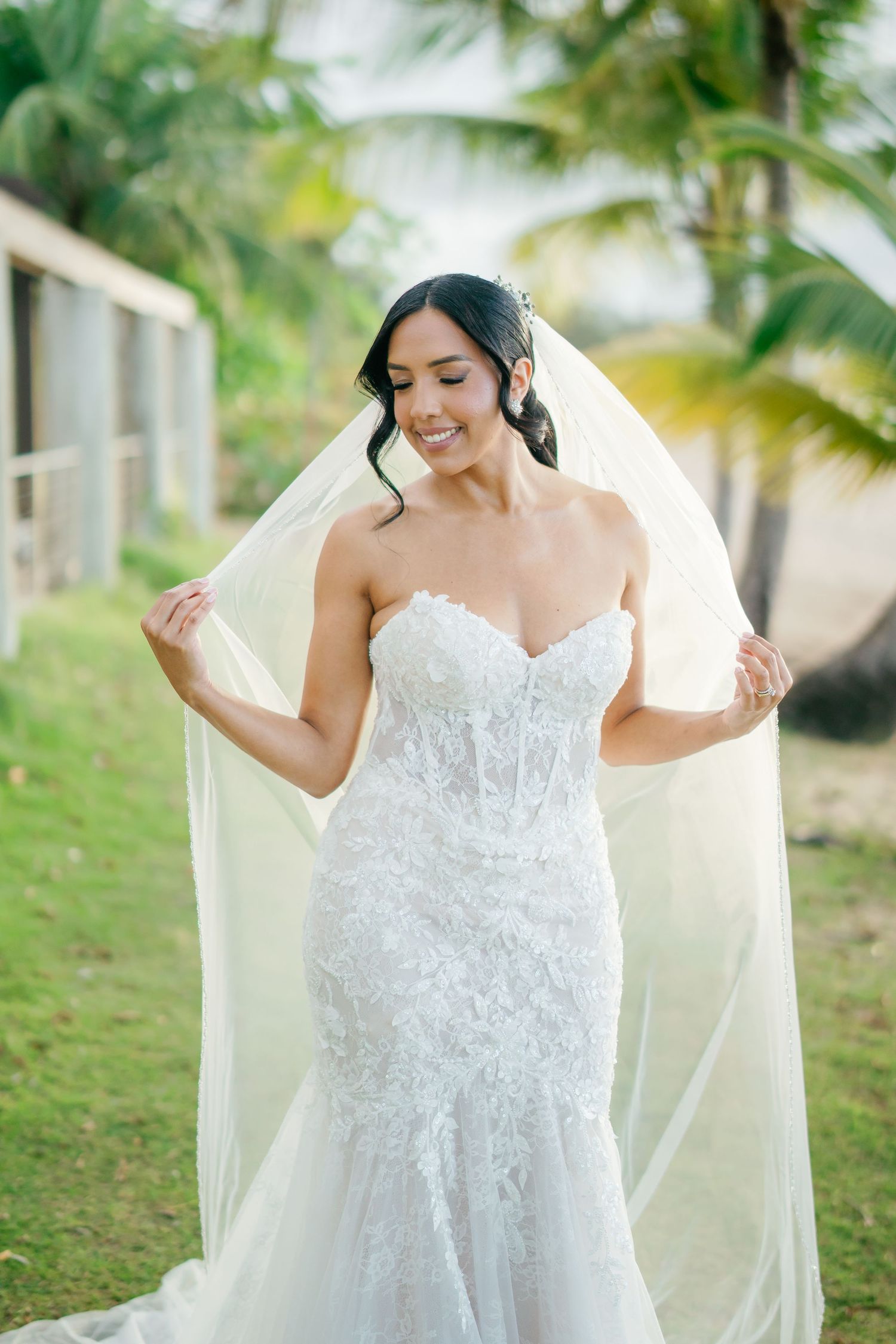 Smiling bride in lace mermaid gown holding flowing veil outdoors with tropical palm trees in background.