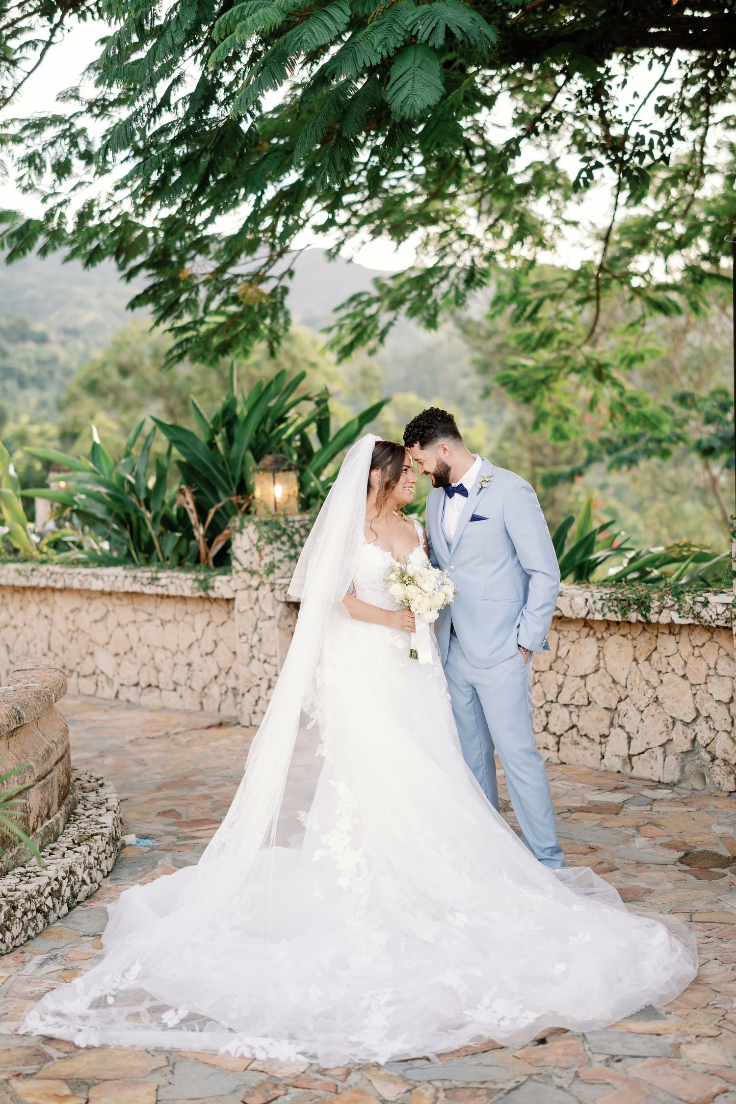 Bride and groom share a romantic moment outdoors, bride in lace gown with veil, groom in light blue suit, lush greenery backdrop.