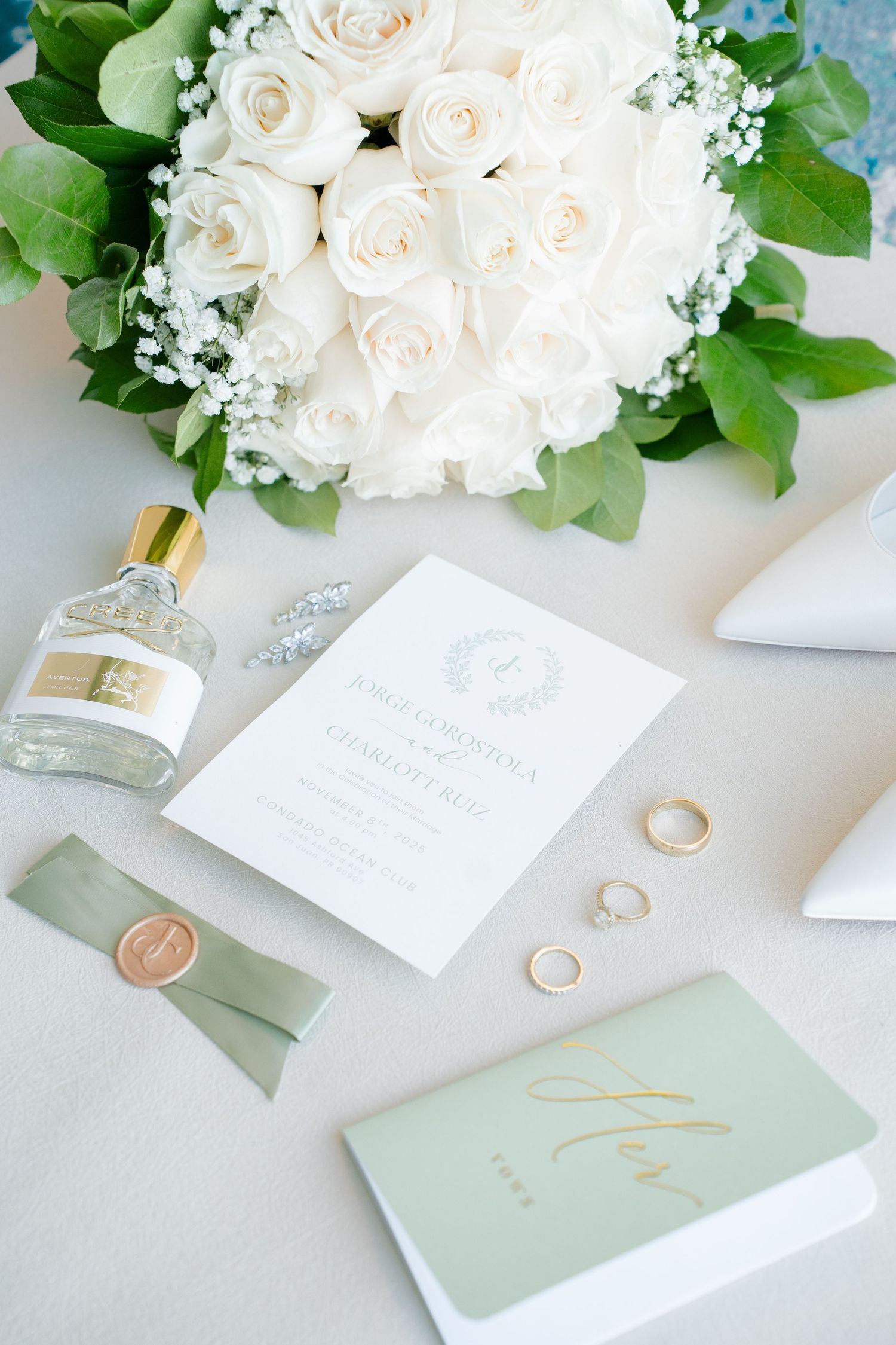 Elegant wedding flat lay with white rose bouquet, gold rings, perfume bottle, sage green invitation suite, and wax seal ribbon.