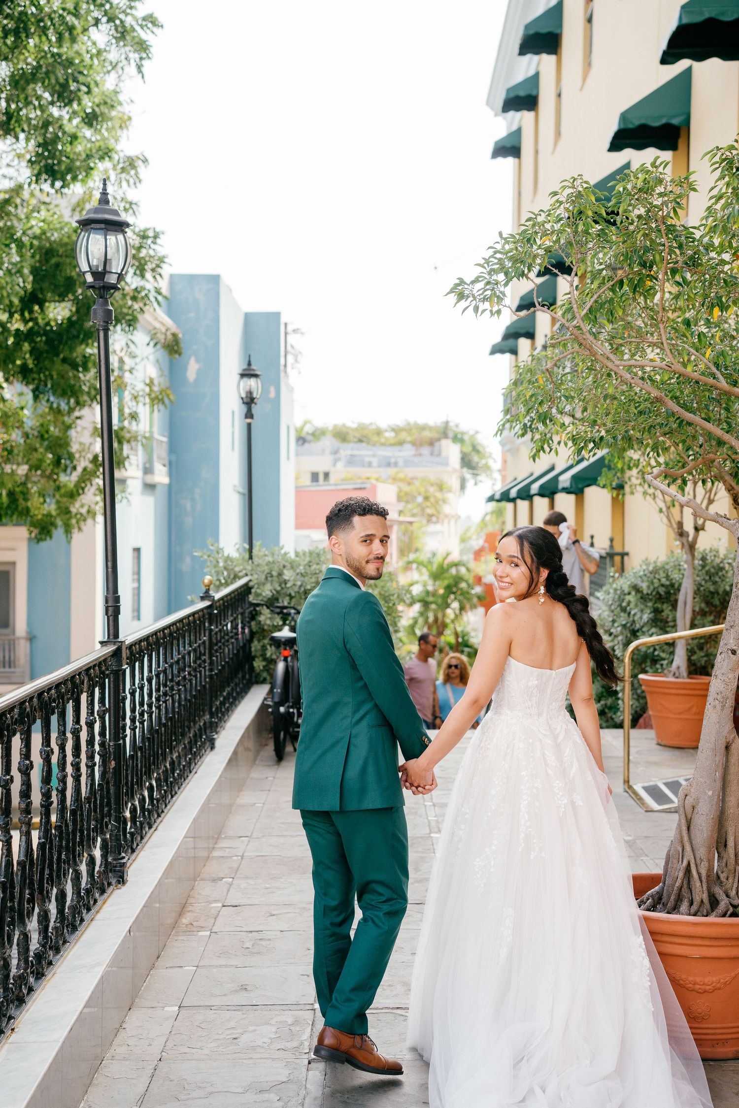Bride and groom holding hands on a colorful balcony walkway, groom in teal suit looks back smiling.
