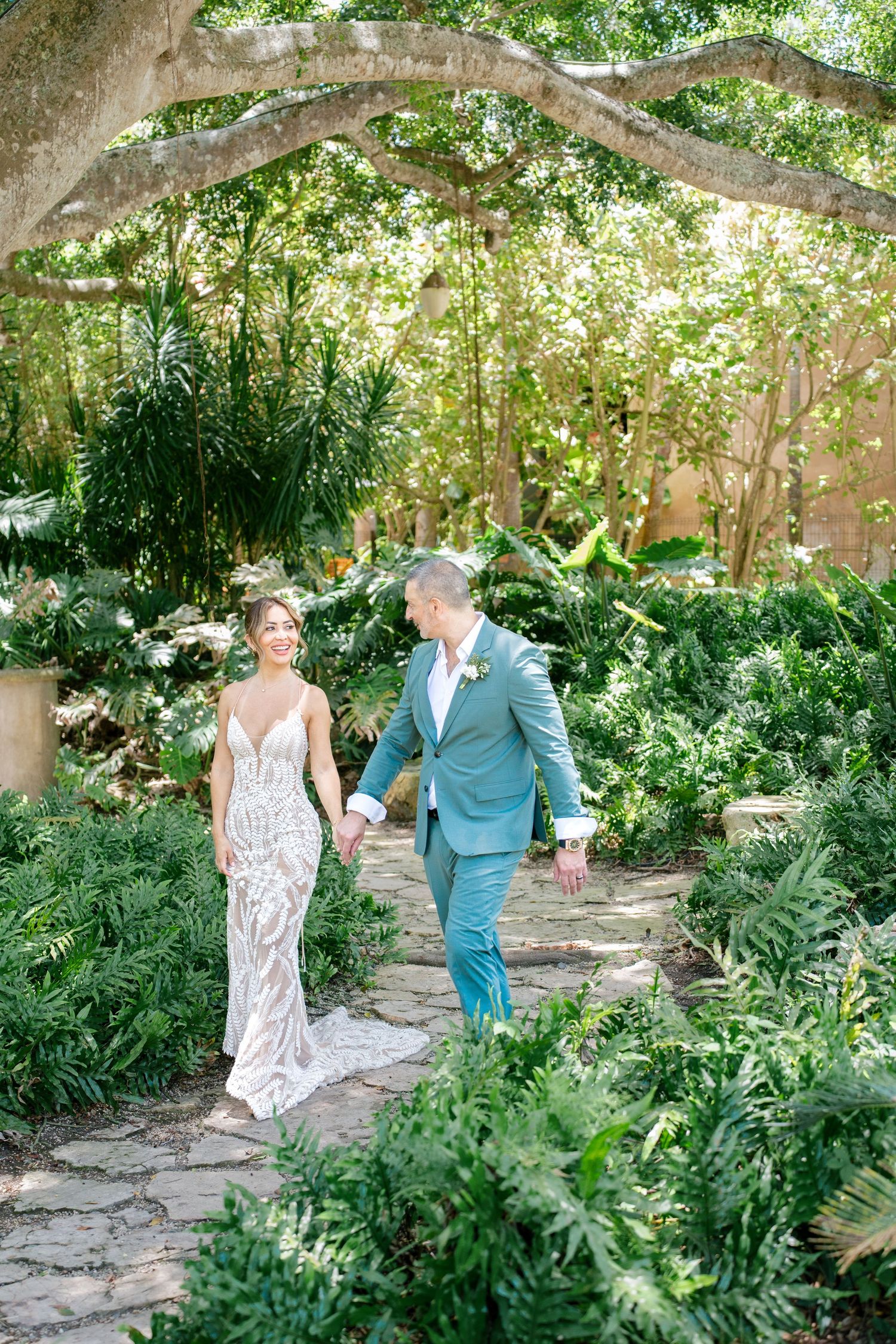 A couple walks hand in hand along a stone path through a lush garden with overhanging tree branches and ferns.