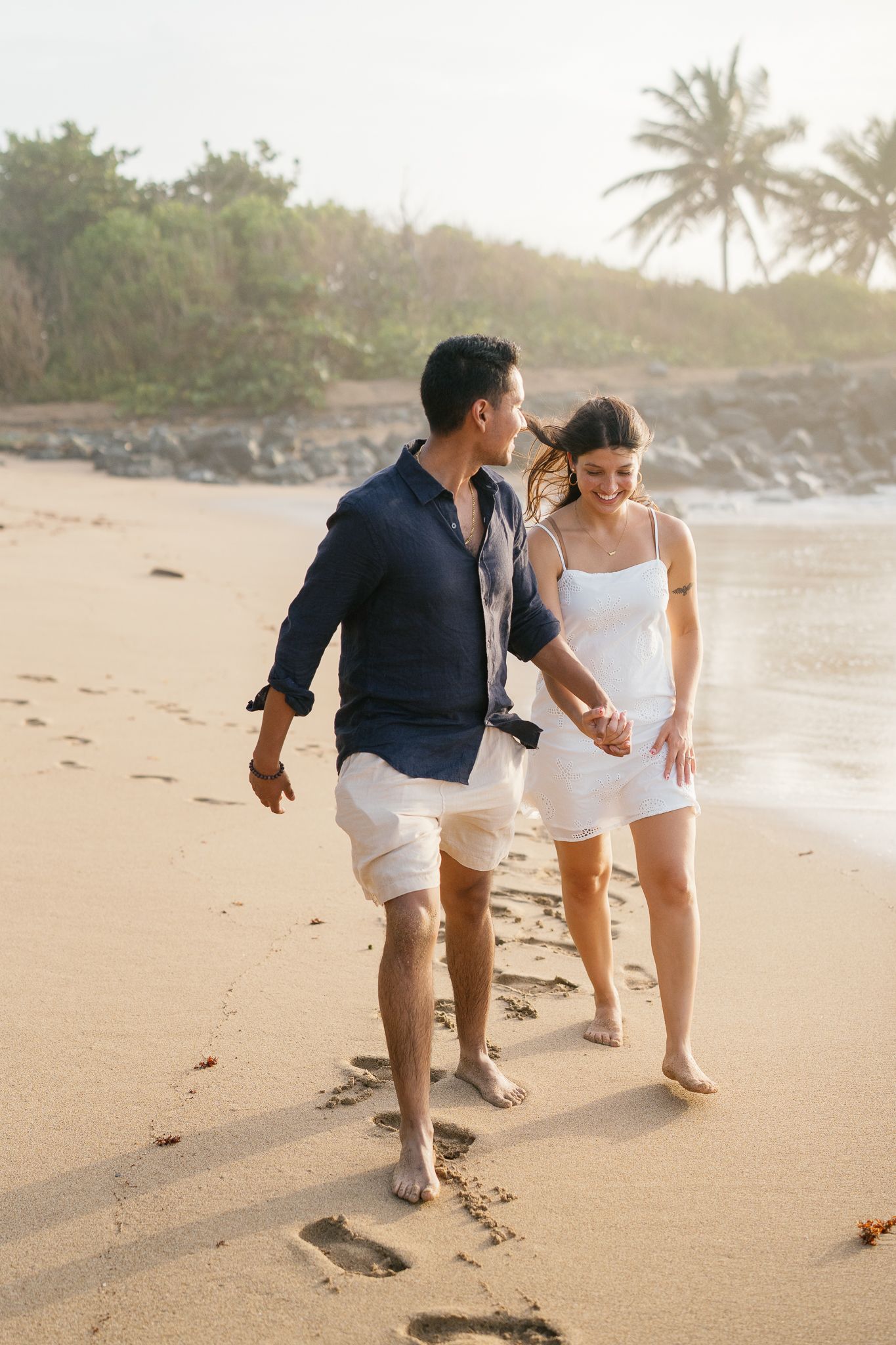 A couple in white outfits walks along a tropical beach at sunset while playing in the sand.