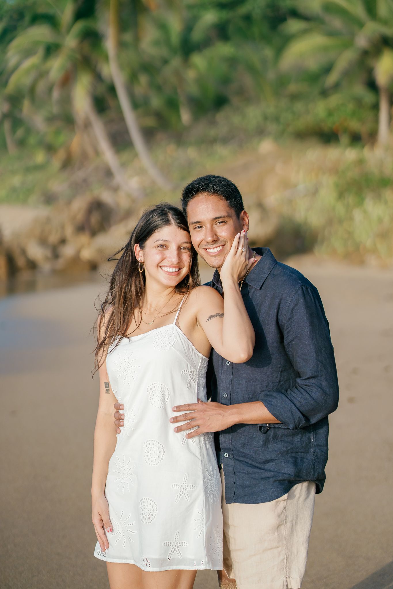 A couple shares a tender moment together on a tropical beach with palm trees in the background.