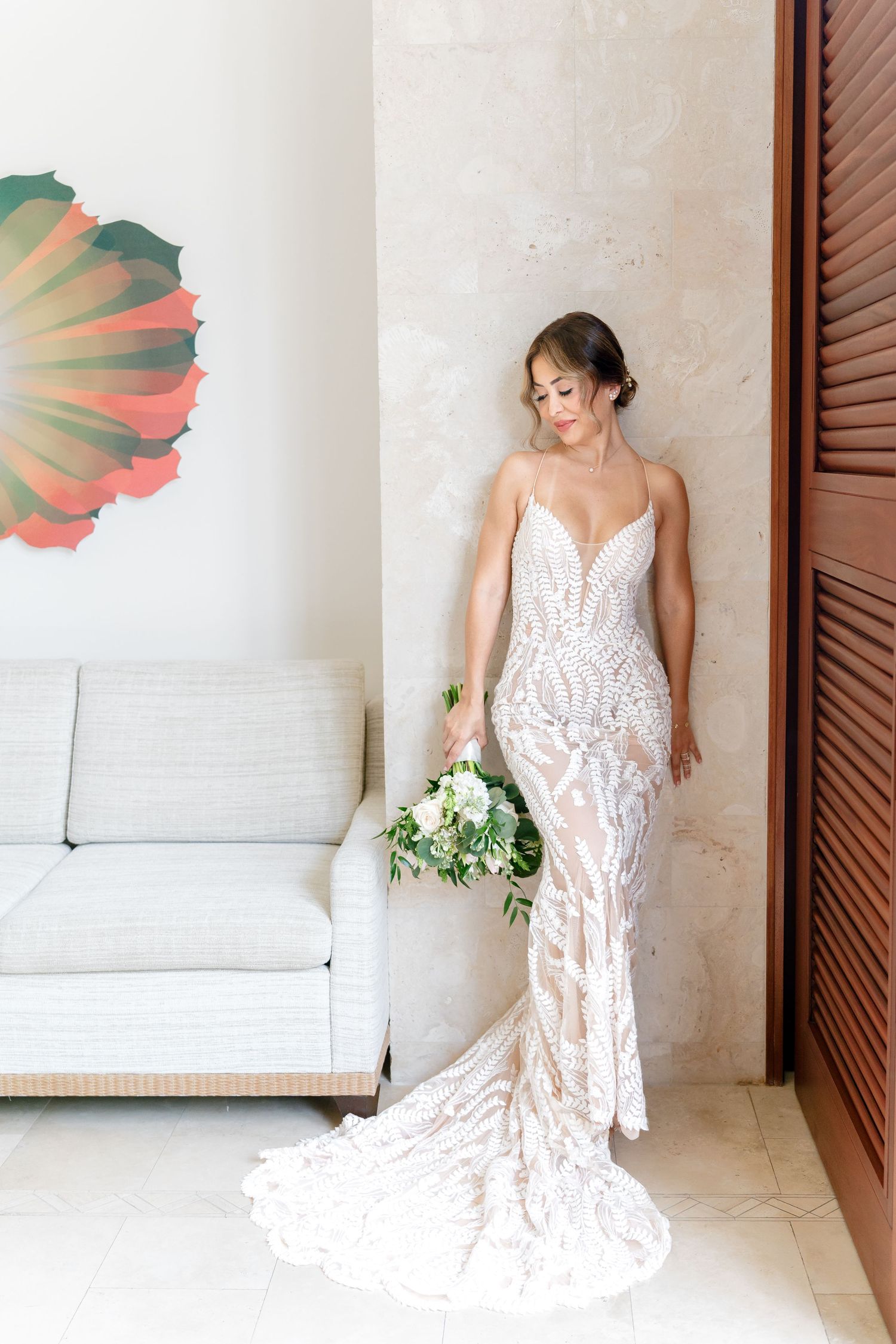An elegant white lace wedding gown with a fitted silhouette and flowing train is displayed in a bright interior space.
