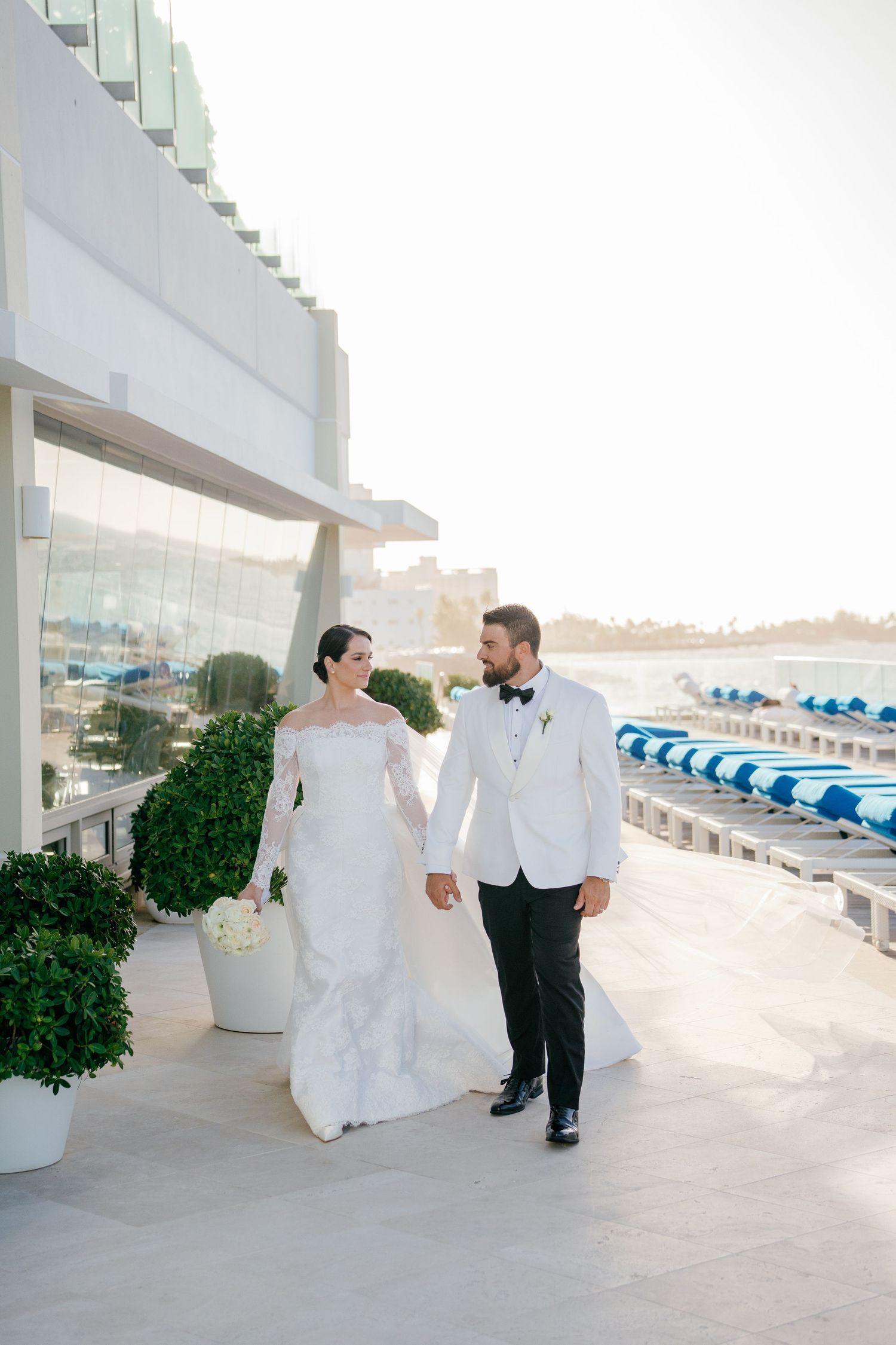 Bride and groom walk hand-in-hand on a sunlit waterfront terrace, she in a lace gown, he in a white tuxedo jacket.