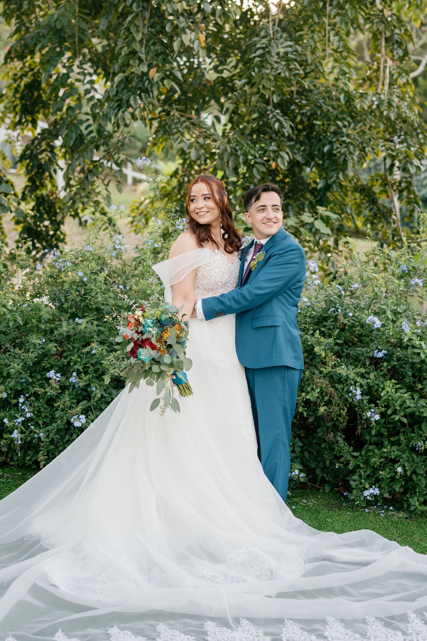 Bride in white gown holding colorful bouquet with groom in blue suit embracing in lush garden setting.