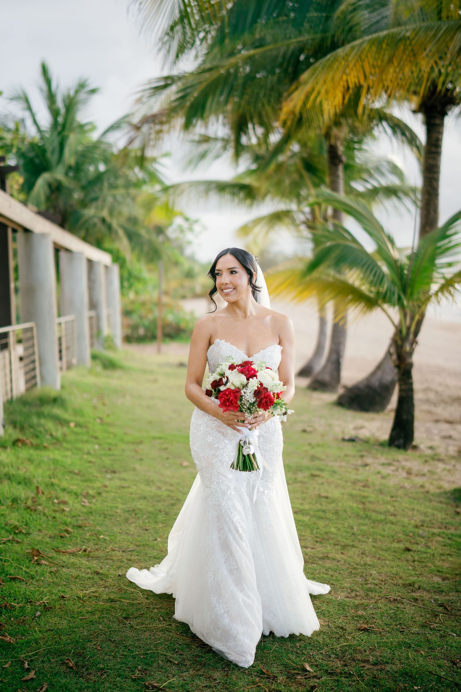 Bride in white lace mermaid gown holding red and white bouquet standing among tropical palm trees on beach.