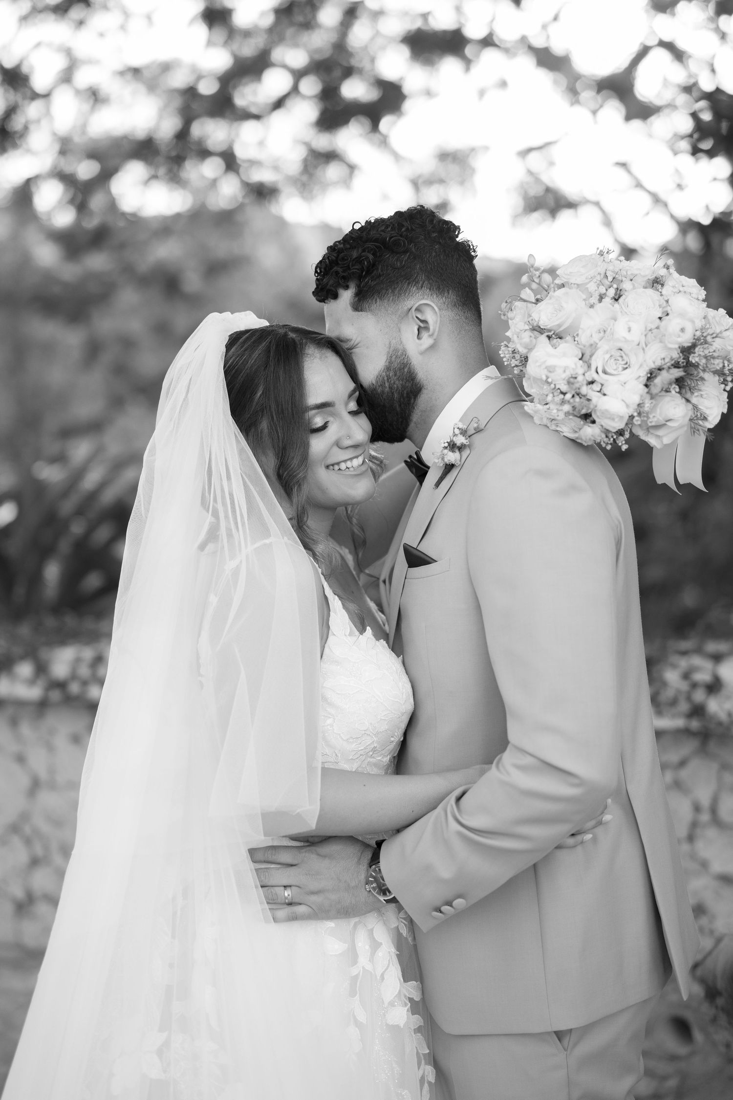 Black and white wedding photo of bride in lace gown and veil smiling as groom in tan suit kisses her cheek outdoors.