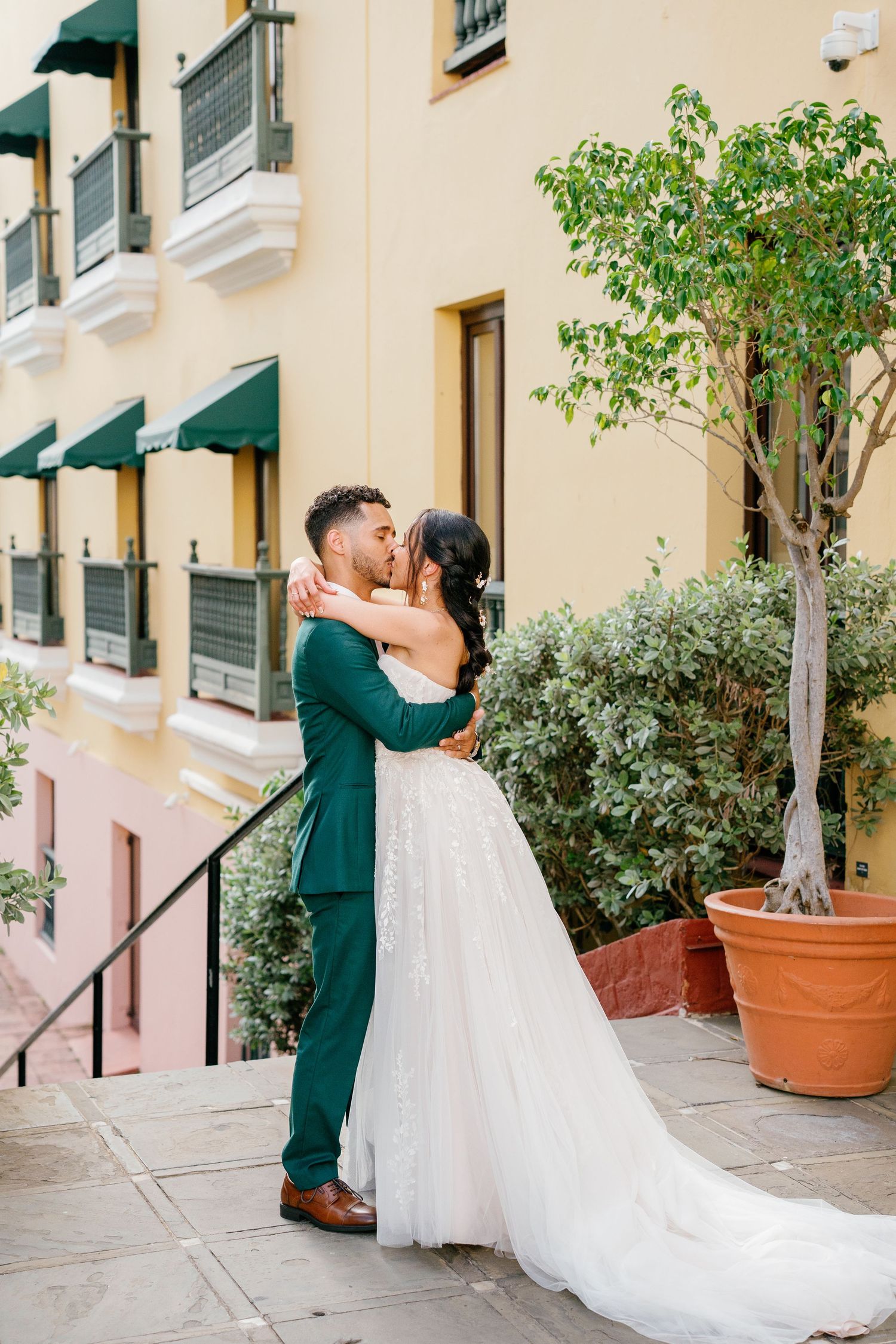 Bride and groom share a romantic kiss in front of a yellow building, groom in green suit, bride in white gown.