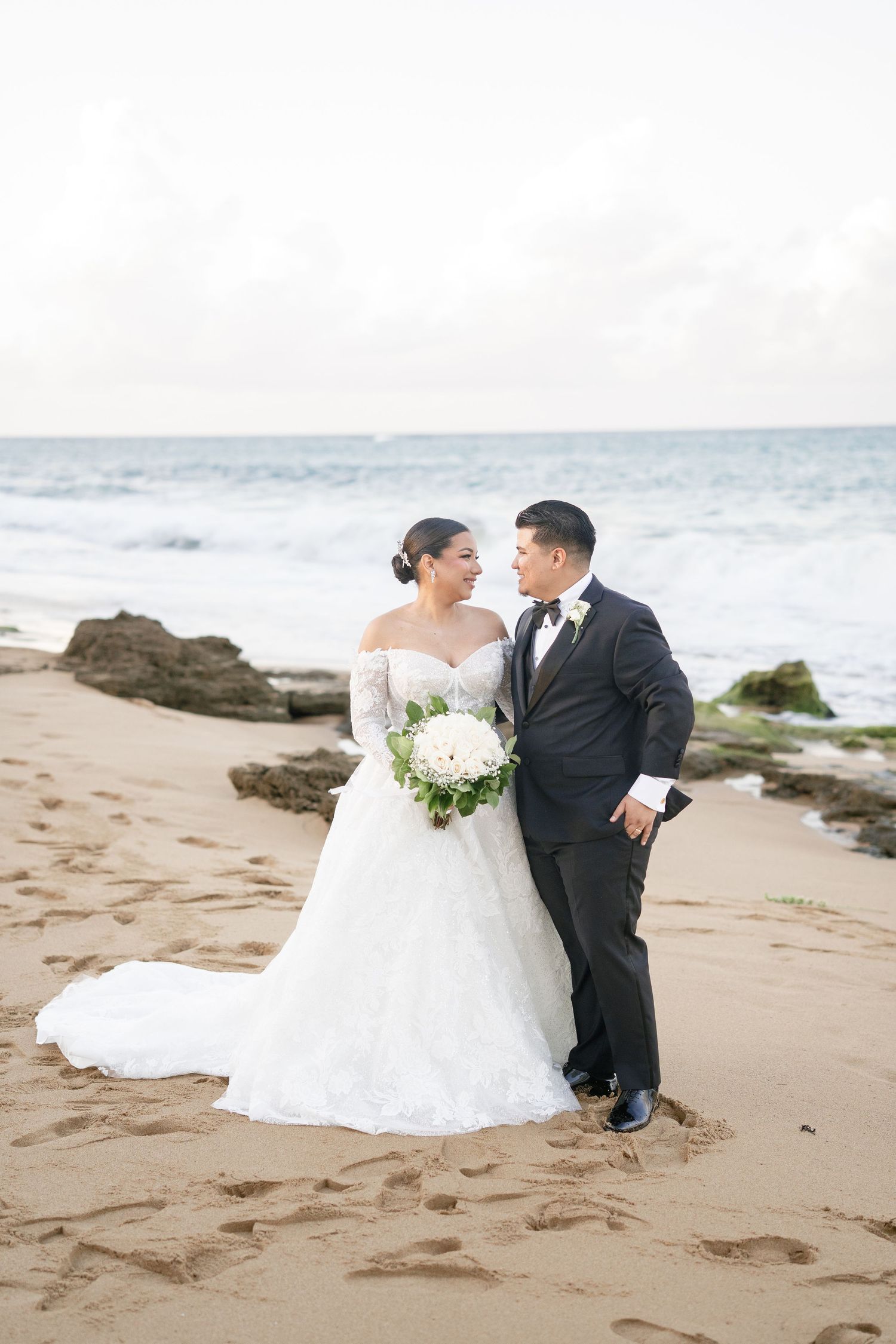 Bride and groom share a romantic moment on a sandy beach, she holds white bouquet in lace gown, he wears black tuxedo.