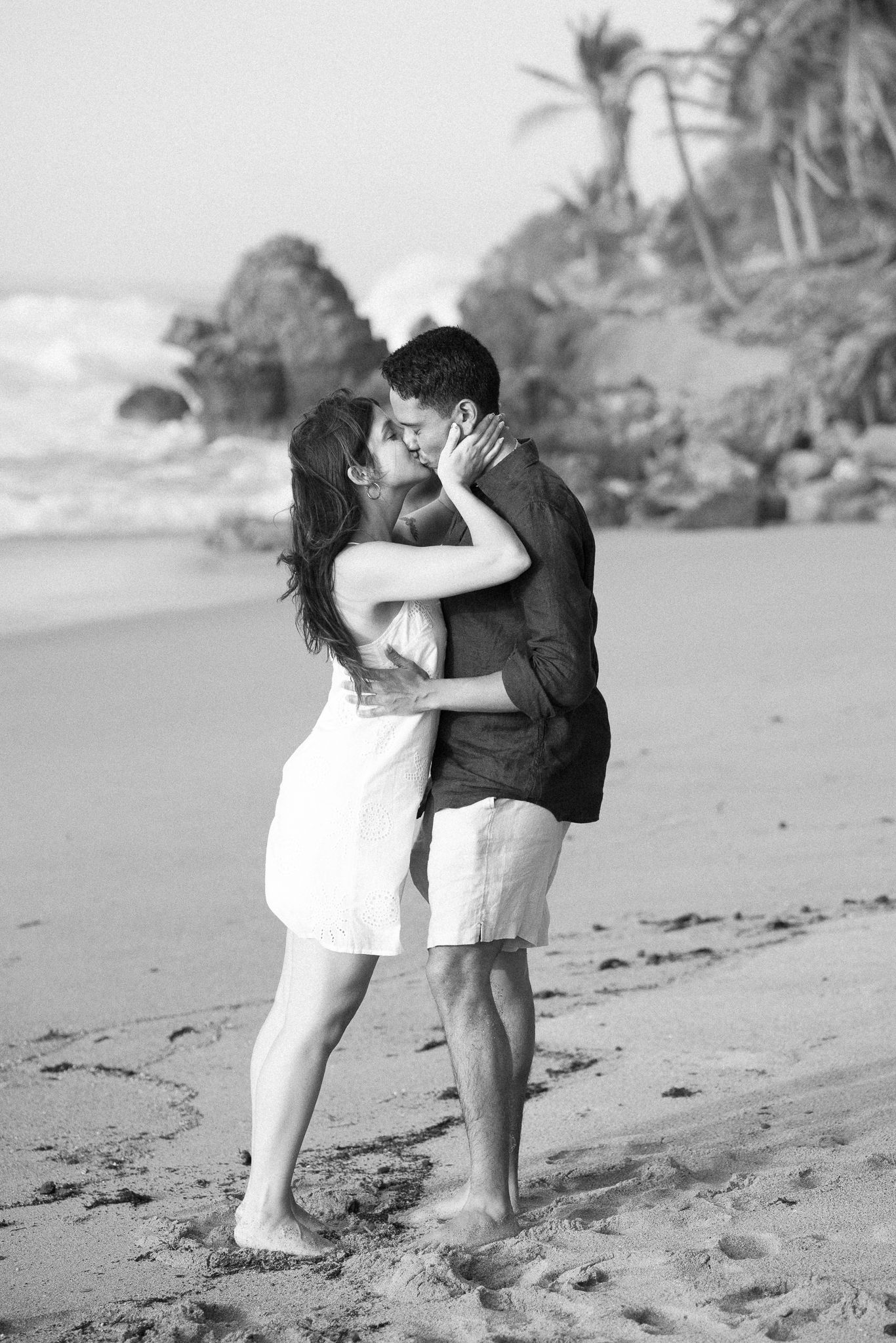 Black and white photo series of two children sharing a sweet kiss on a sandy beach with palm trees in the background.