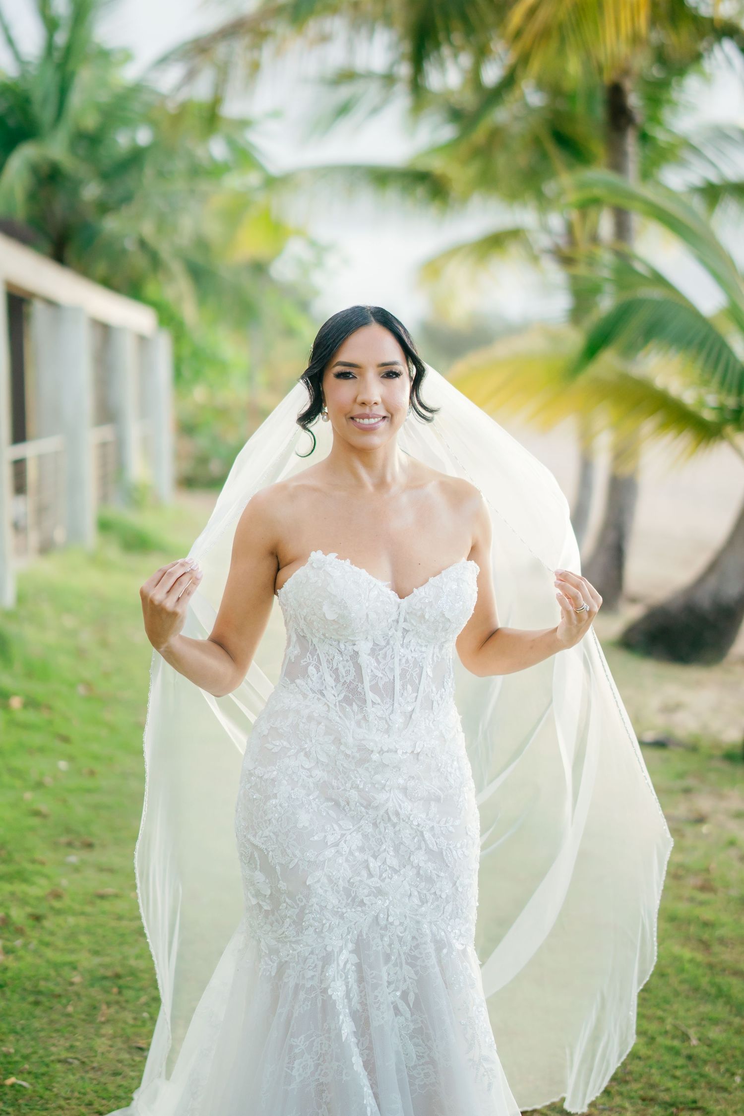 Smiling bride in strapless lace mermaid gown holding flowing veil outdoors with tropical palm trees background.