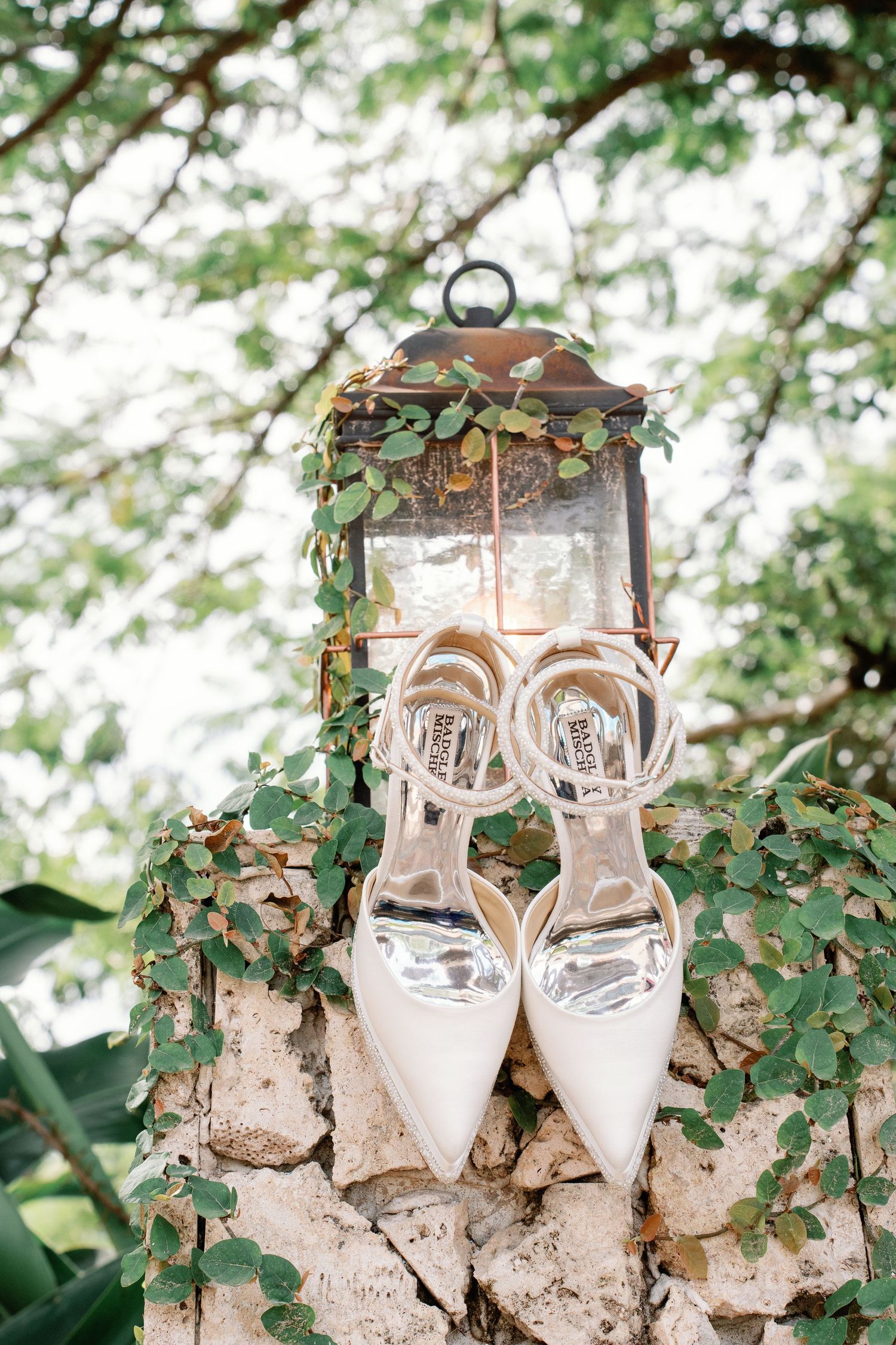 Elegant white Badgley Mischka bridal heels displayed on a stone wall beside a vintage lantern with green ivy.