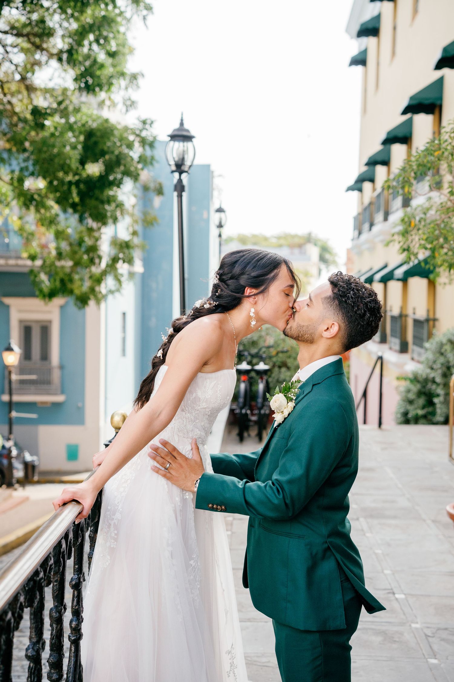 A bride and groom share a romantic kiss on a balcony, dressed in a white gown and emerald green suit.