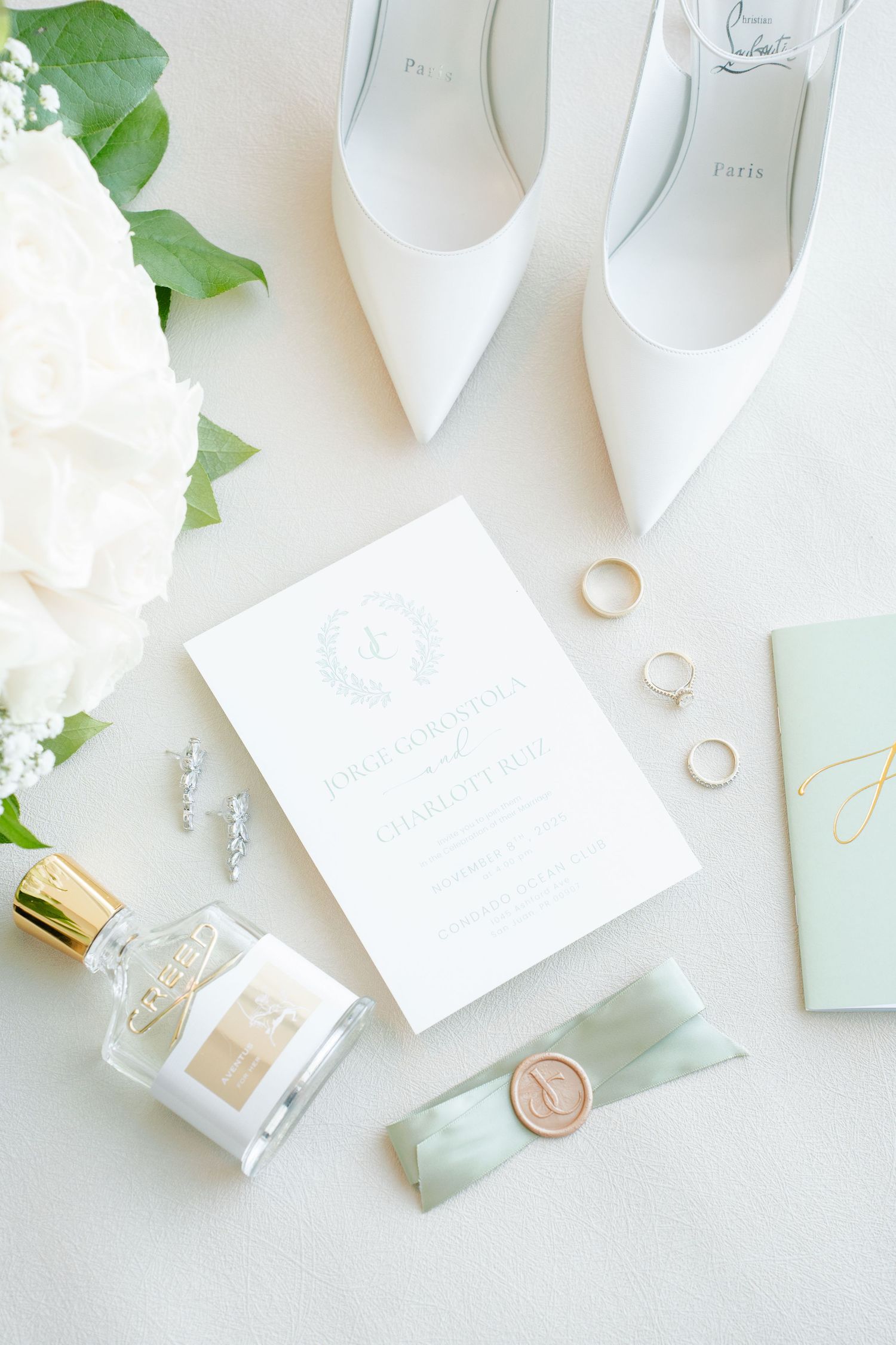 Elegant wedding flat lay with white heels, floral bouquet, gold rings, perfume, wax seal, and a classic wedding invitation.