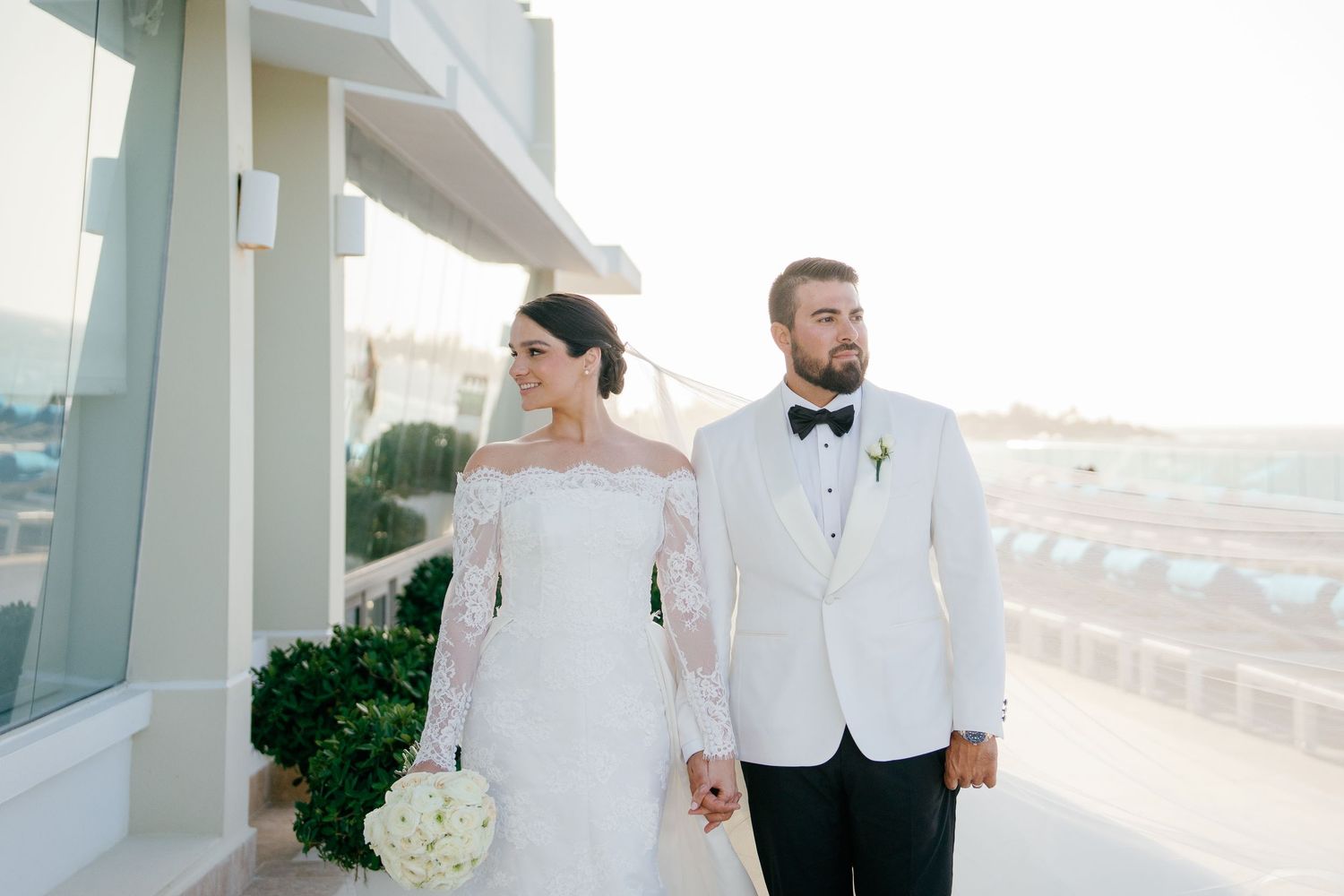 Bride and groom hold hands on a rooftop terrace, she in lace off-shoulder gown, he in white tuxedo with bow tie.