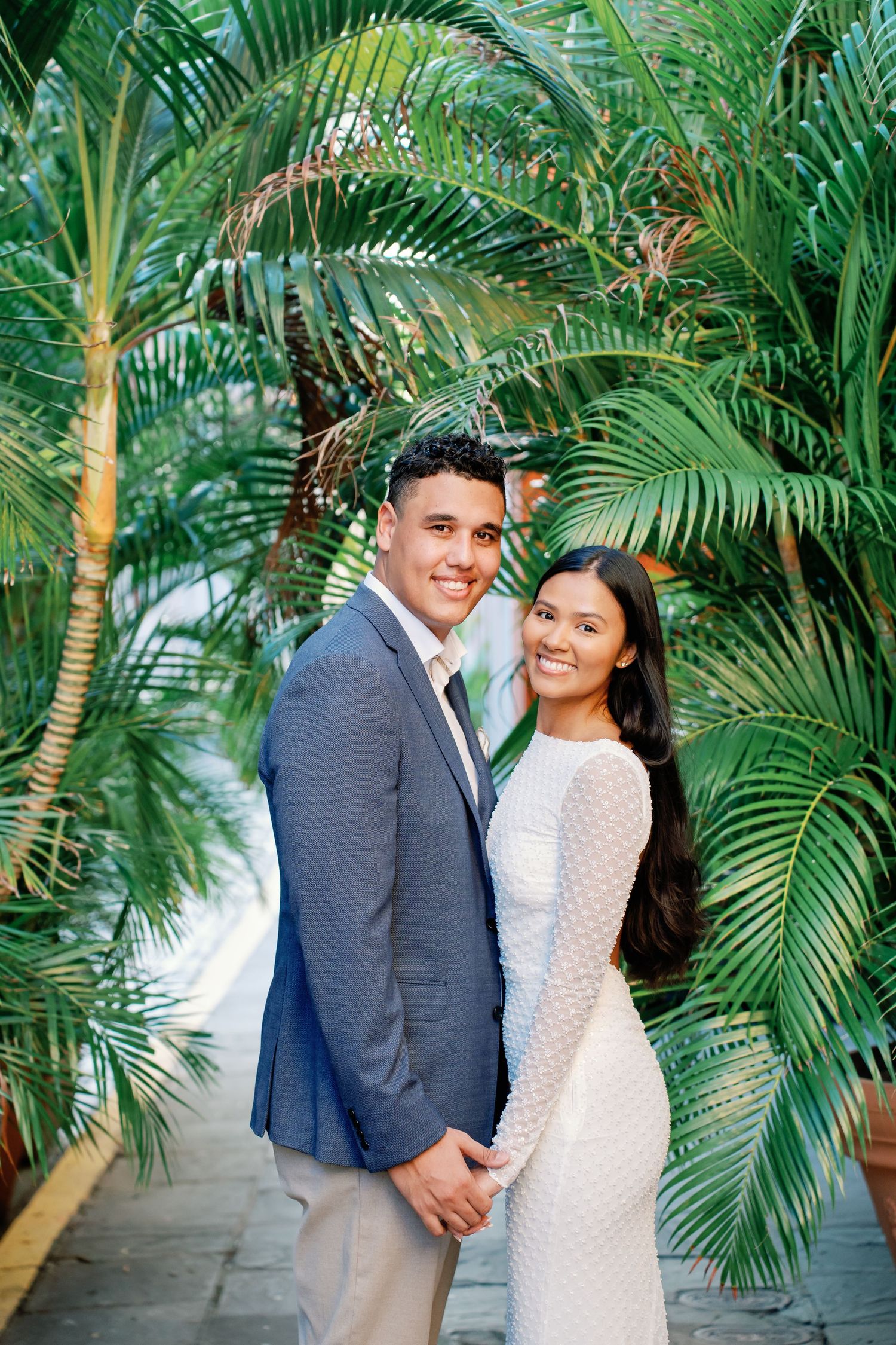 Smiling couple in formal attire posing together among lush tropical palm trees in an elegant outdoor setting.