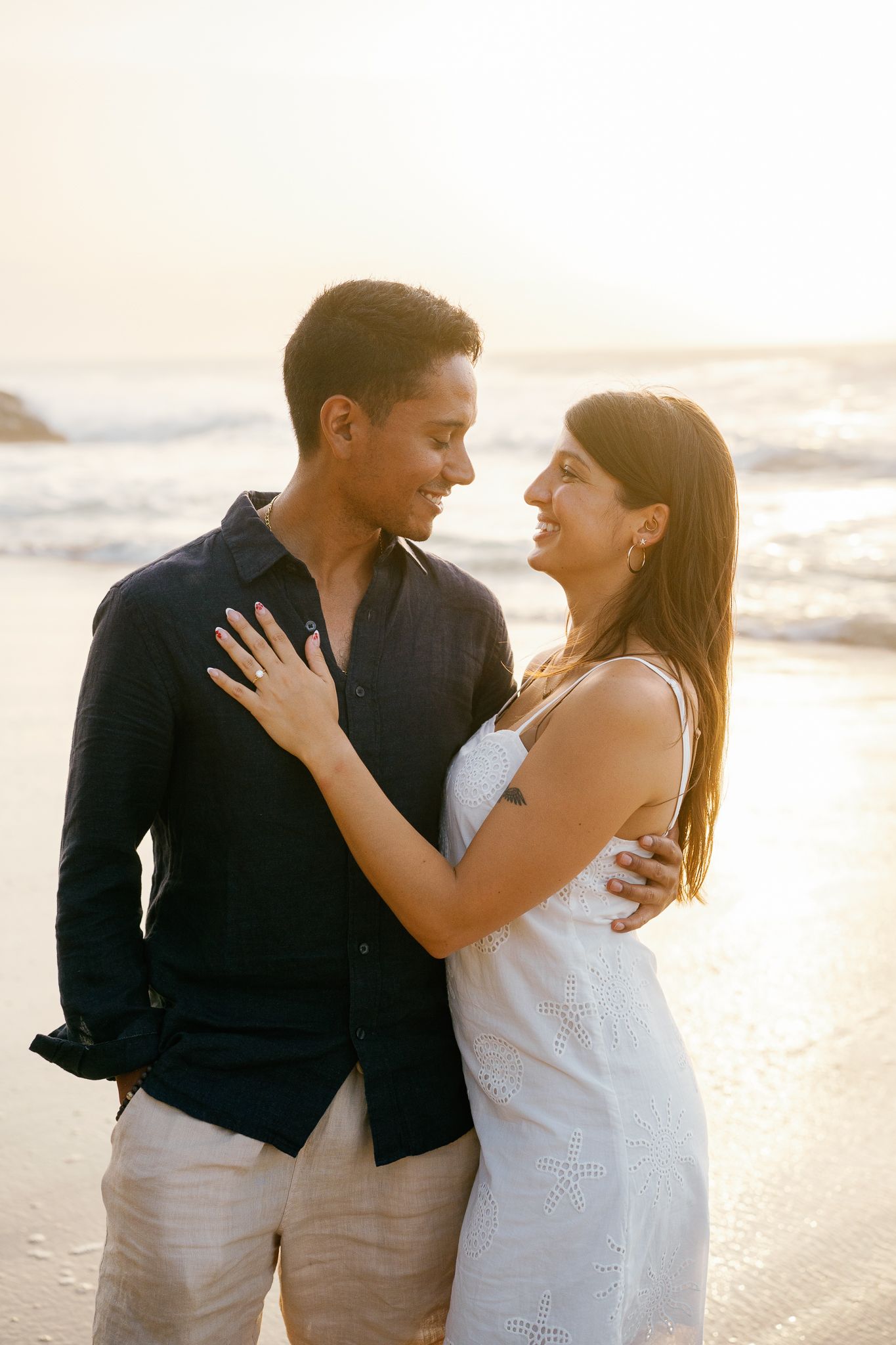 A romantic couple embraces on a sunlit beach during sunset, creating a series of intimate moments in white and black attire.