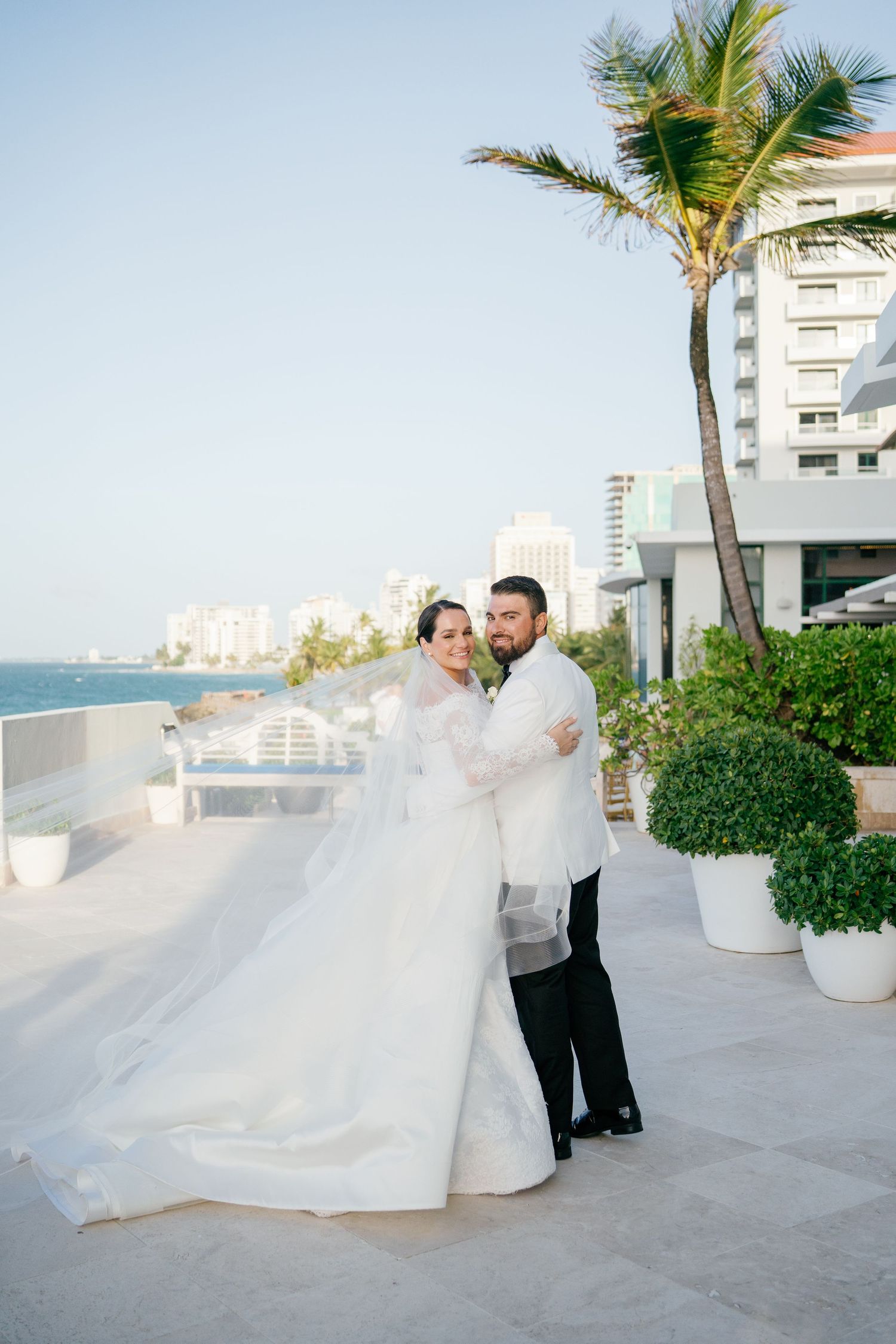 Bride and groom posing on a rooftop terrace with palm trees and ocean views on their wedding day.