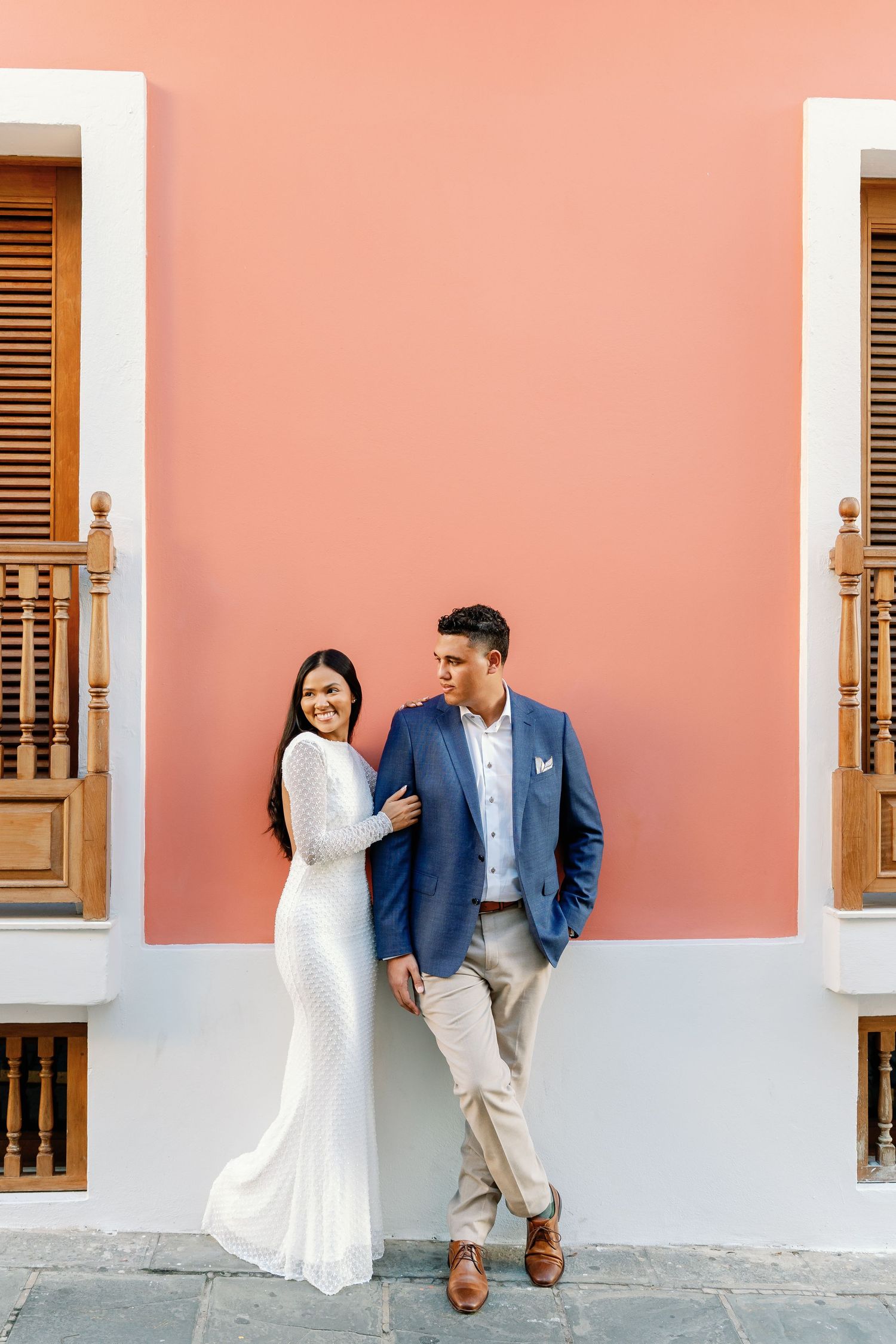Elegant couple posing against a coral pink wall with wooden balconies in a romantic urban setting.