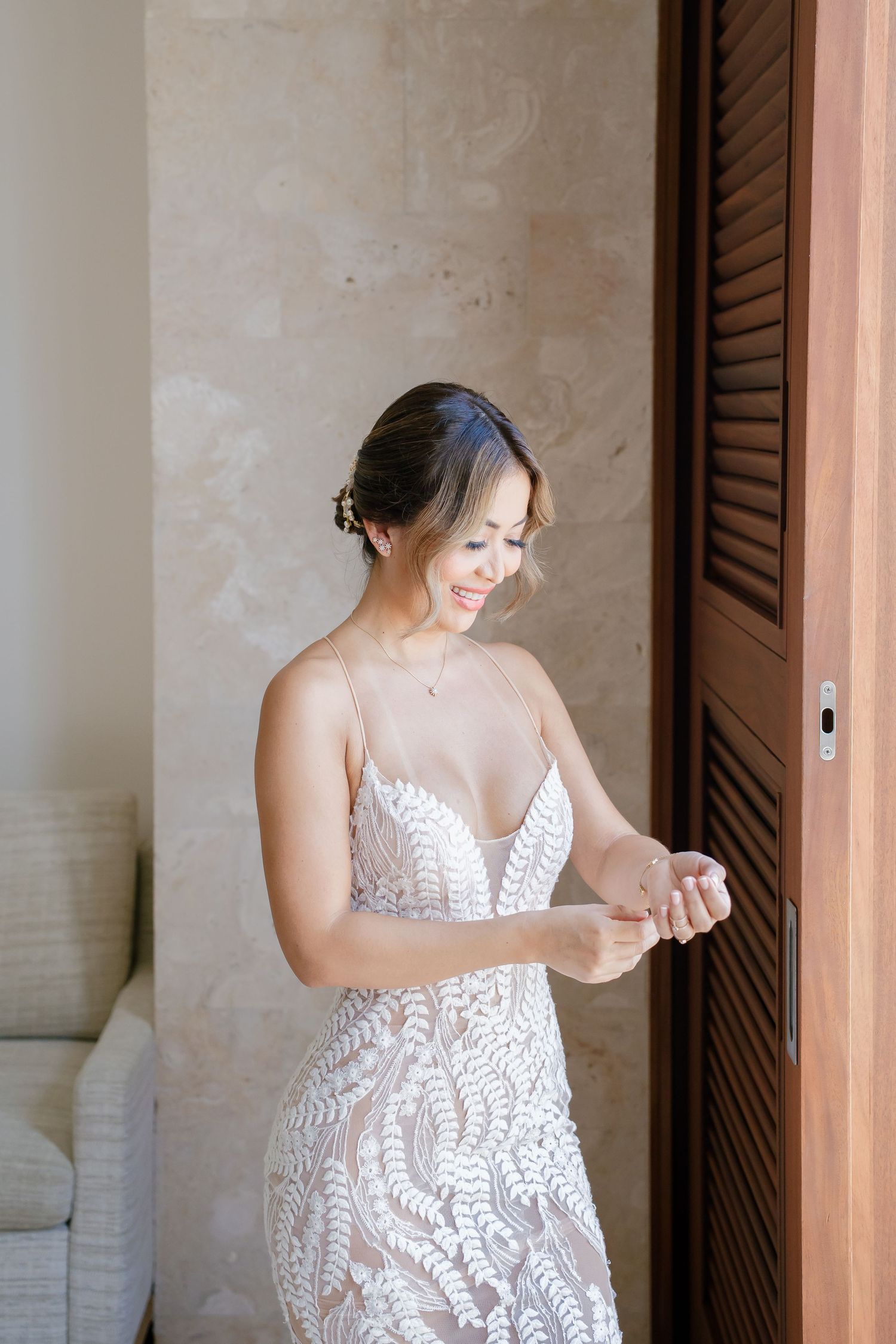 A bride in an elegant lace wedding dress adjusts her jewelry near wooden shutters in a soft-lit room.