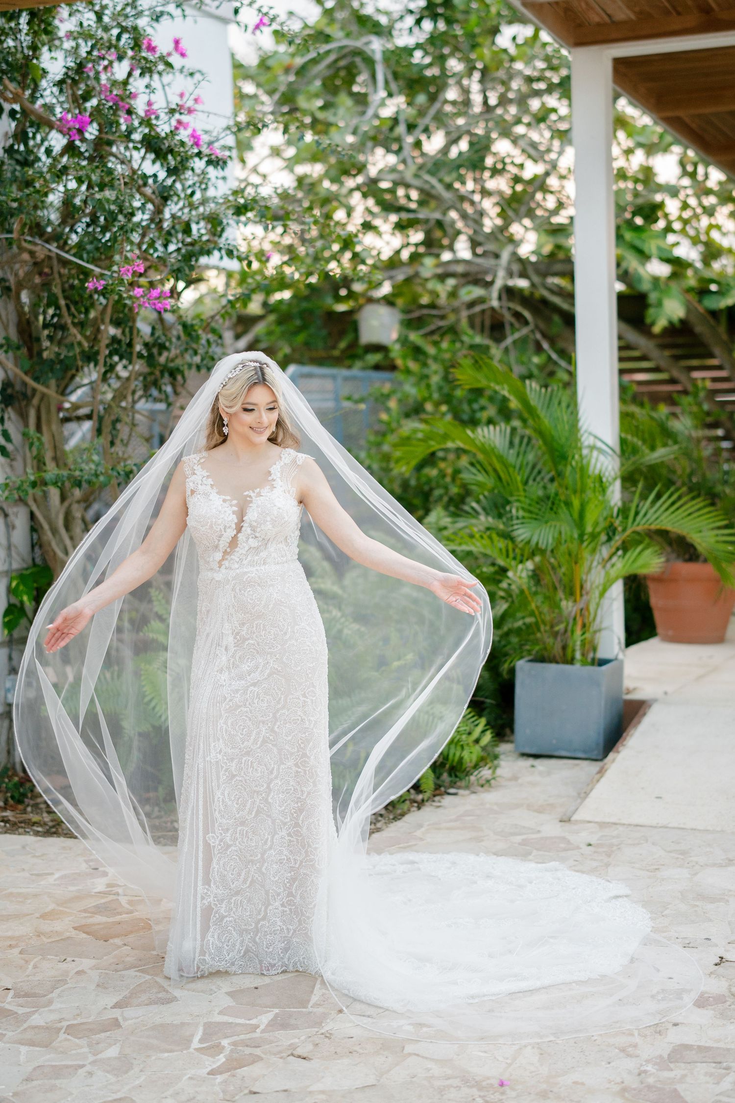 Bride in lace wedding gown and flowing veil poses outdoors among tropical greenery on stone patio.