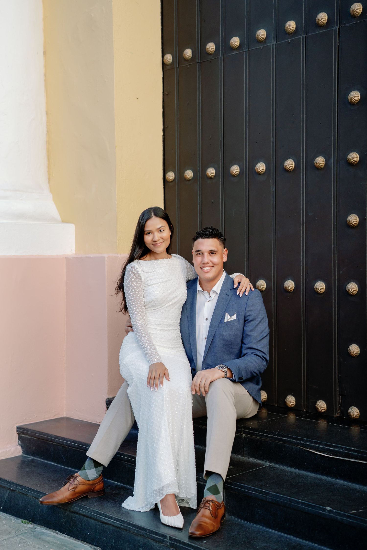 Couple in wedding attire sitting on steps in front of ornate black door, woman in white dress, man in blue suit.