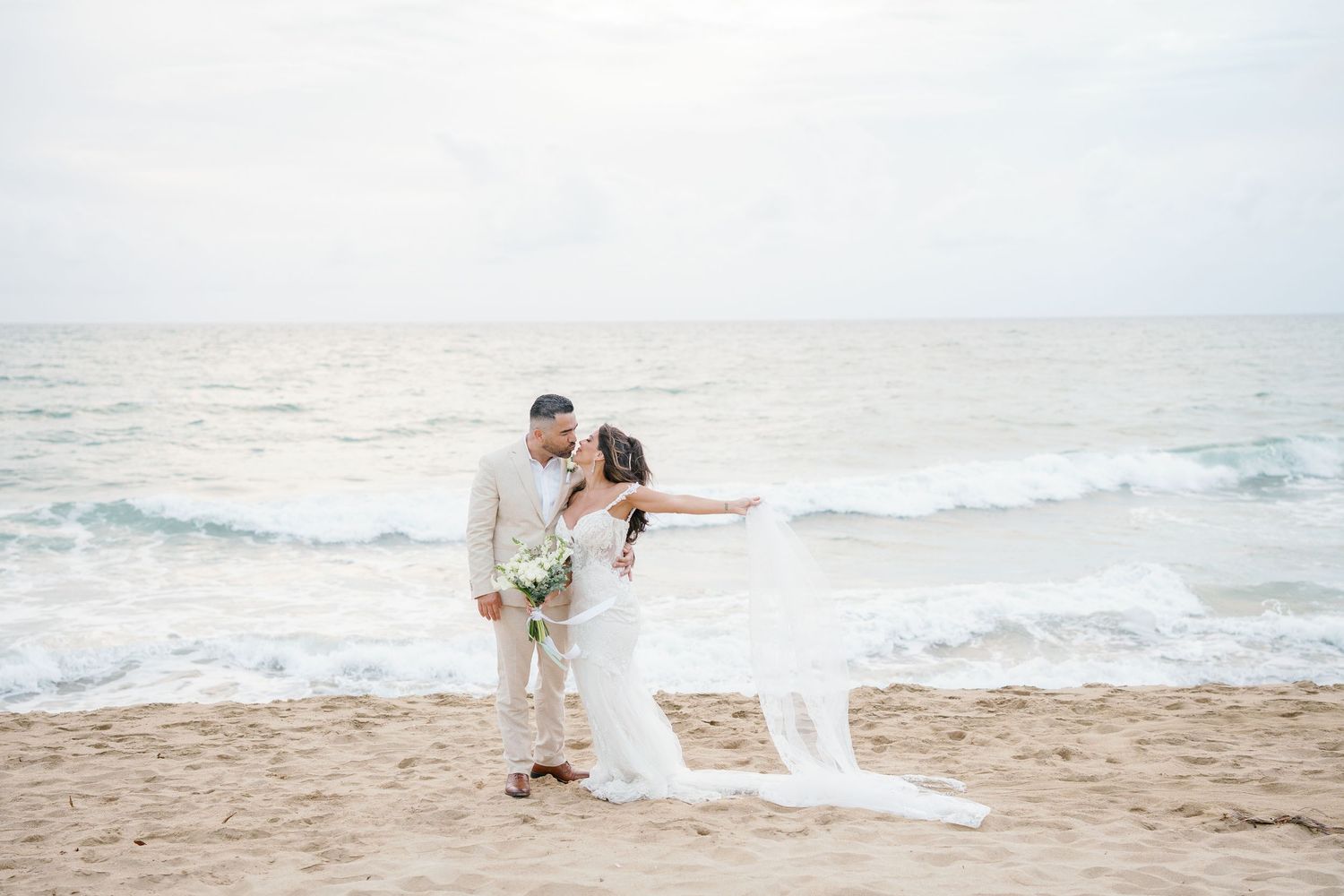 Couple in wedding attire poses on sandy beach with ocean waves, bride holding bouquet with veil flowing in breeze.