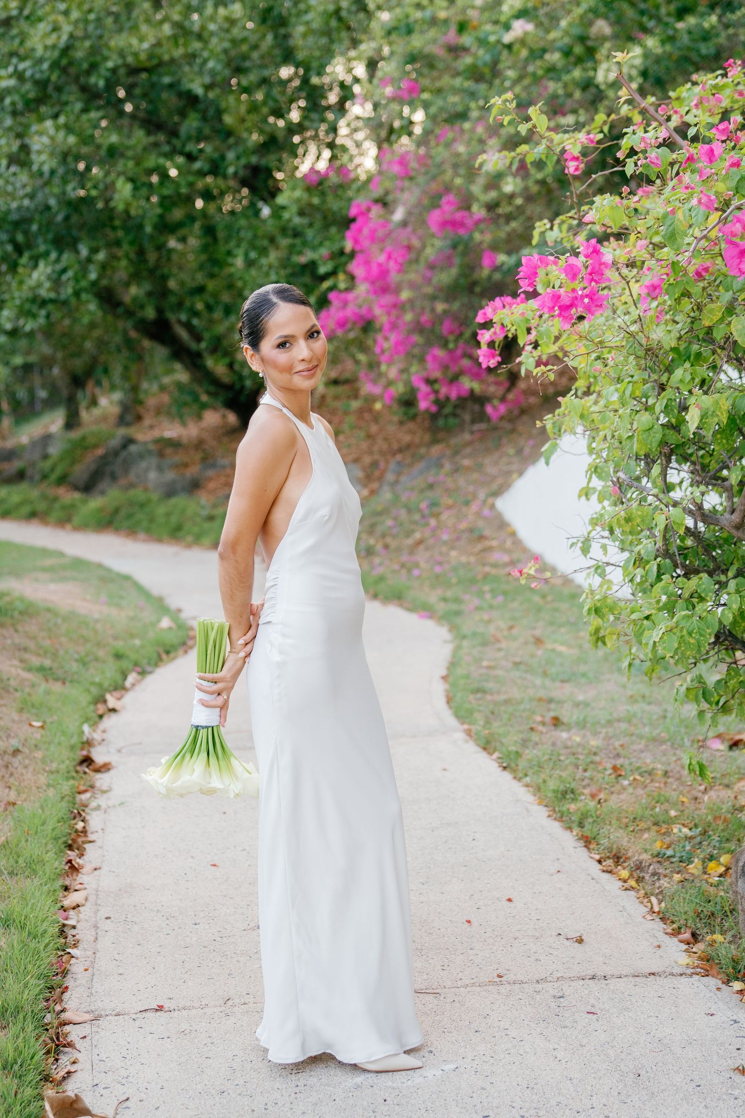 Bride in white sleeveless gown holds calla lily bouquet on garden path with pink bougainvillea blooms.