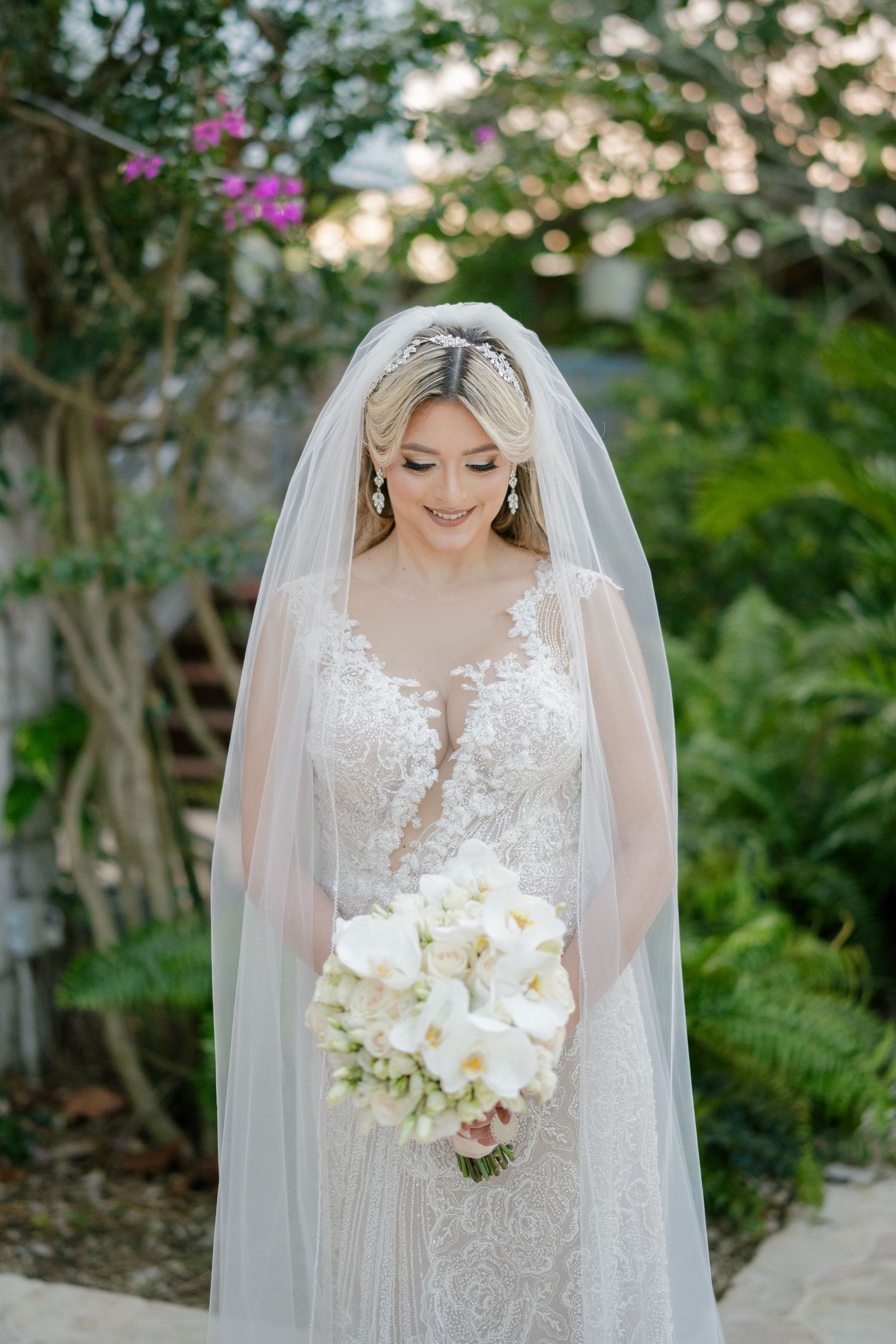 Bride in lace gown and veil holds white orchid bouquet in lush garden setting.