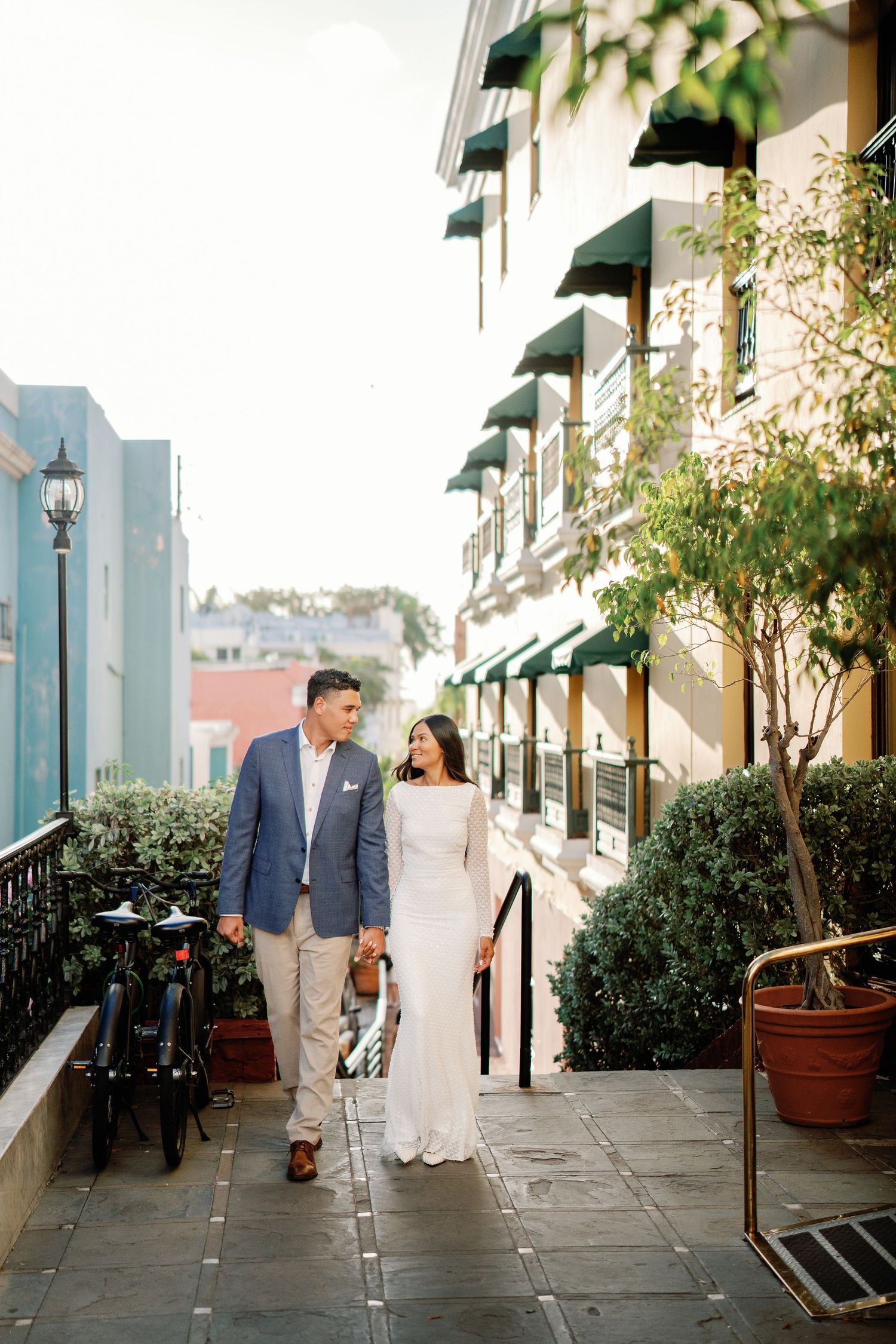 Couple in wedding attire walking hand in hand along a charming outdoor corridor with green awnings.