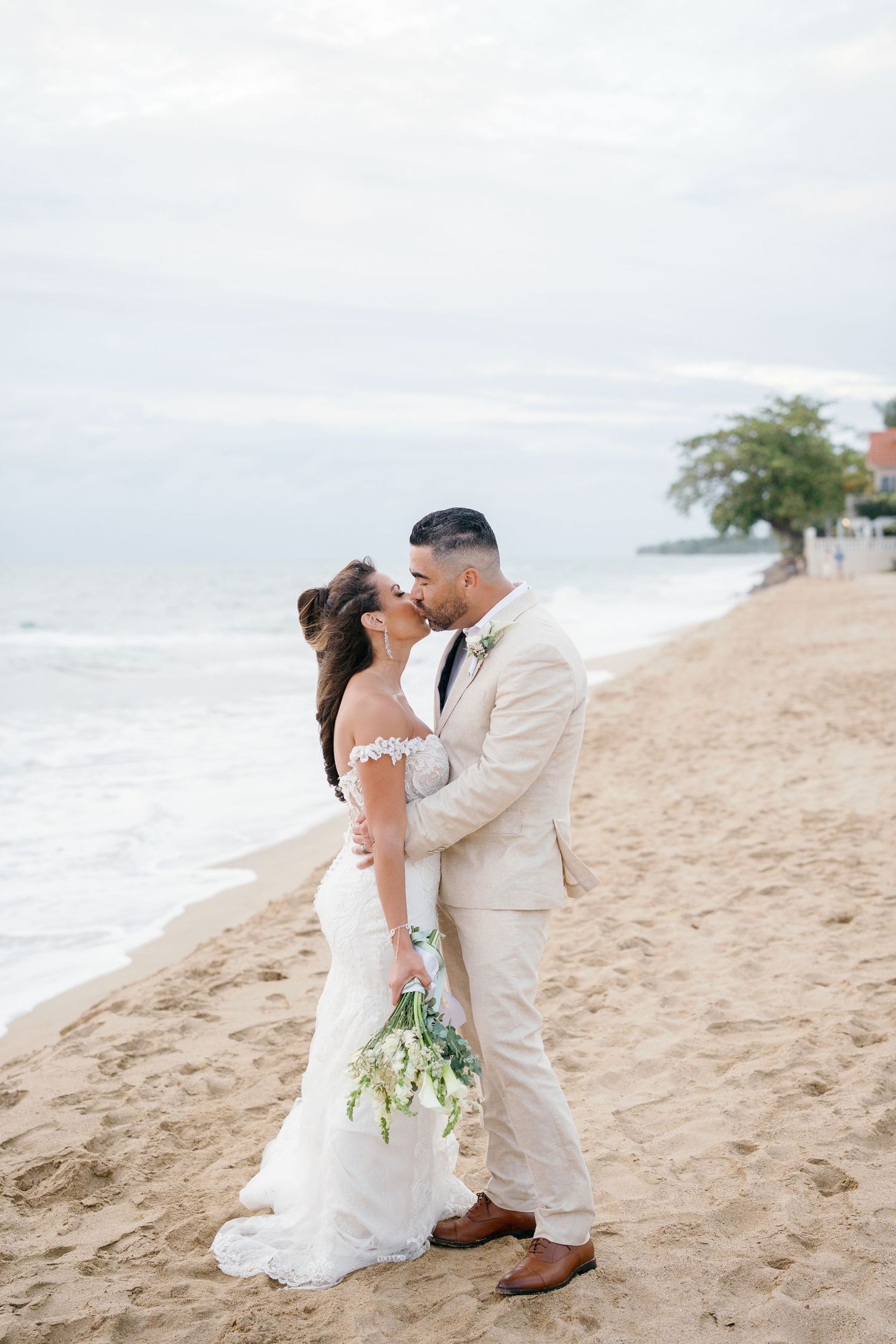 Bride and groom share a romantic kiss on a sandy beach, she in a white gown, he in a beige suit holding her bouquet.