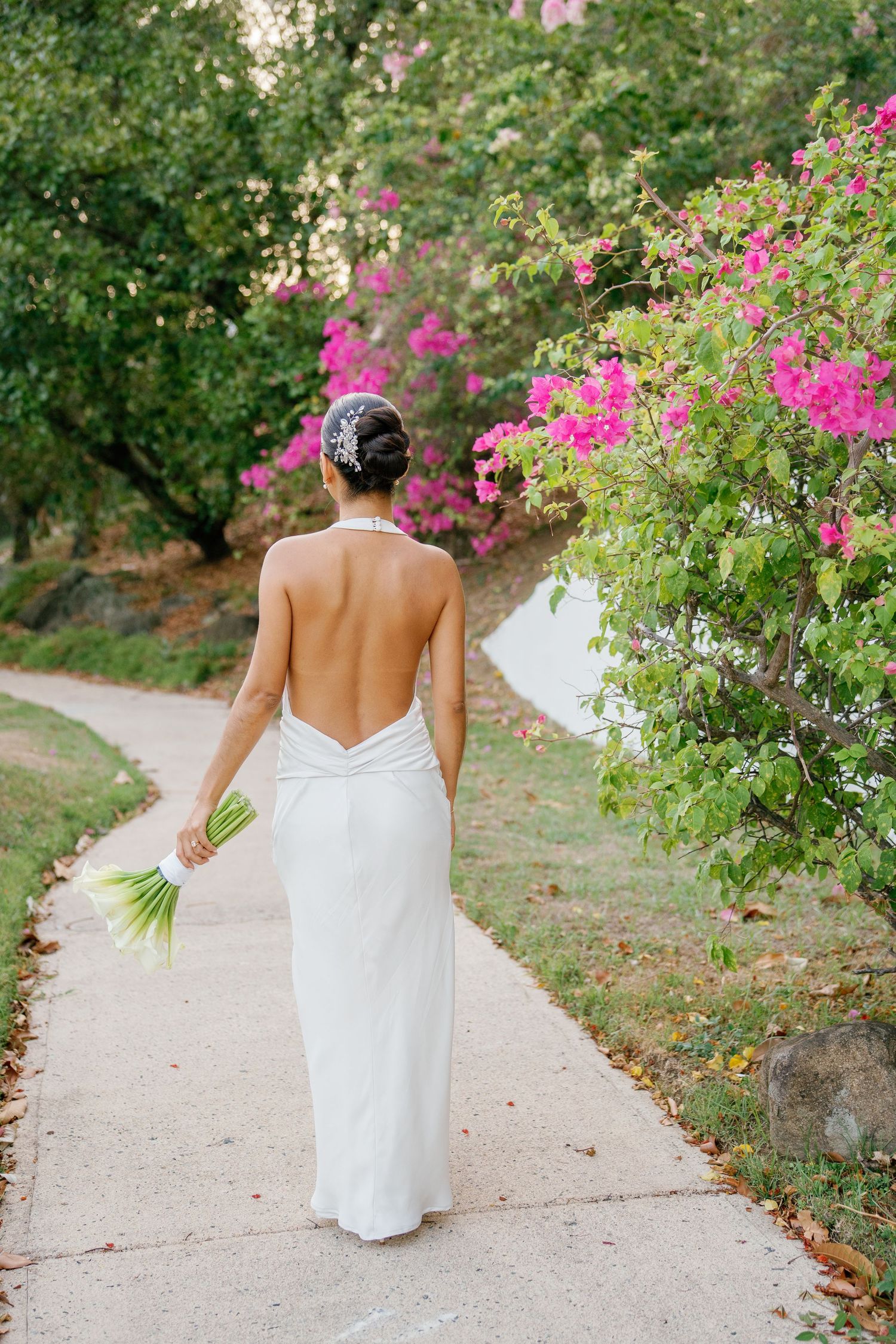 Bride in backless white halter gown walks garden path holding green bouquet near vibrant pink bougainvillea flowers.