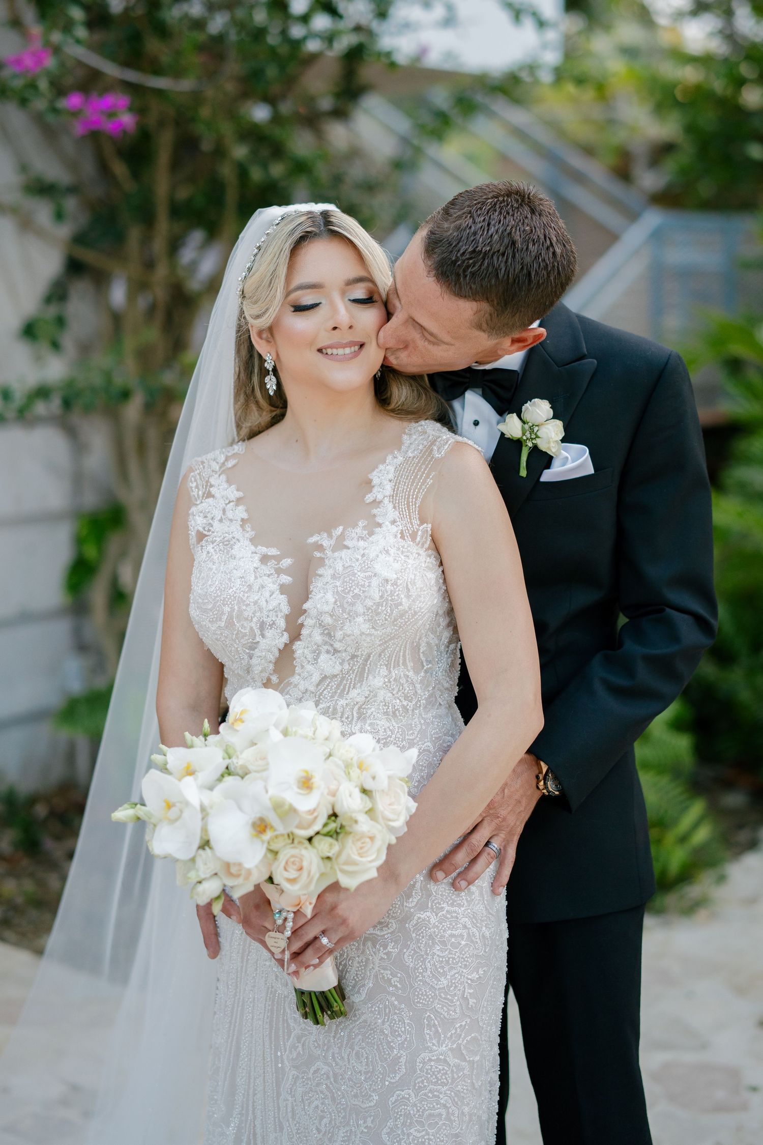 Bride in lace gown holding white orchid bouquet while groom in black tuxedo kisses her cheek outdoors.
