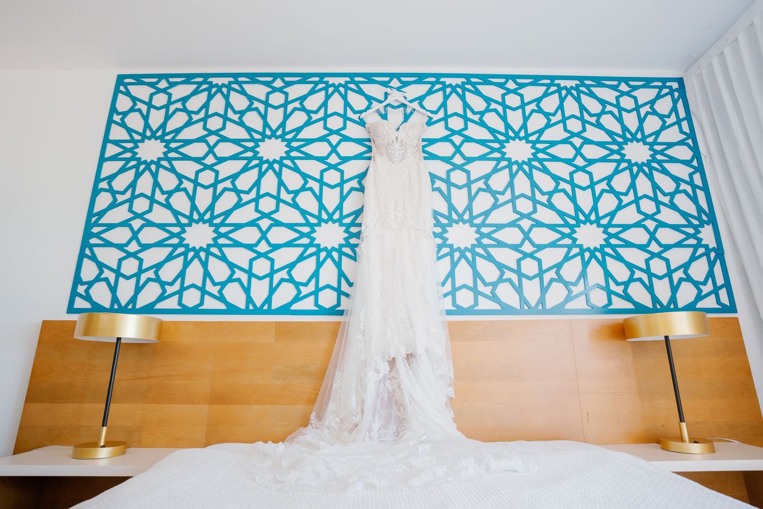 Wedding dress hanging against a turquoise geometric patterned wall above a wooden headboard with gold lamps.