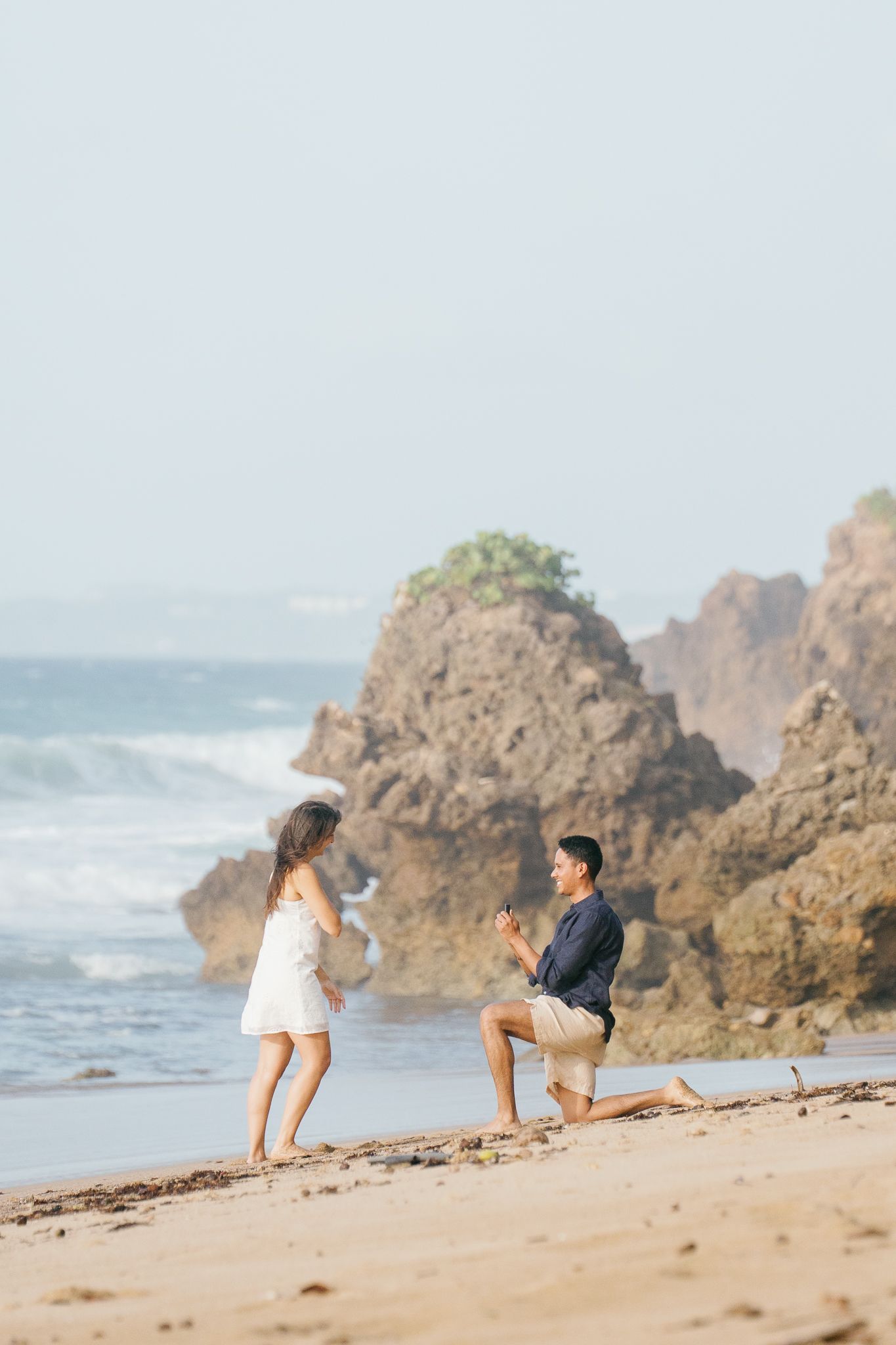 A couple sits together on a sandy beach with dramatic rocky cliffs and crashing waves in the background.