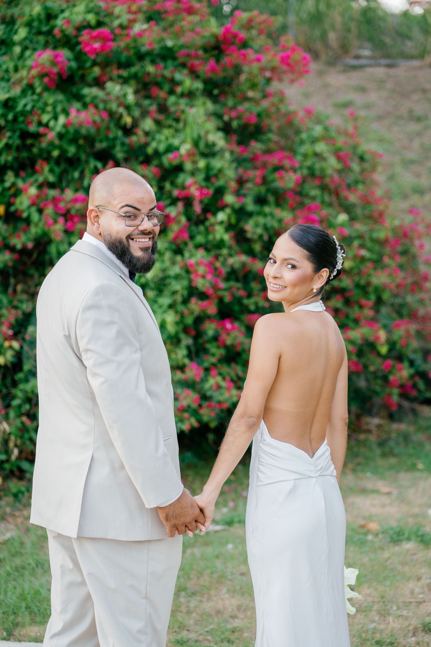 Smiling bride in backless white gown and groom in cream suit hold hands before vibrant pink bougainvillea blooms.