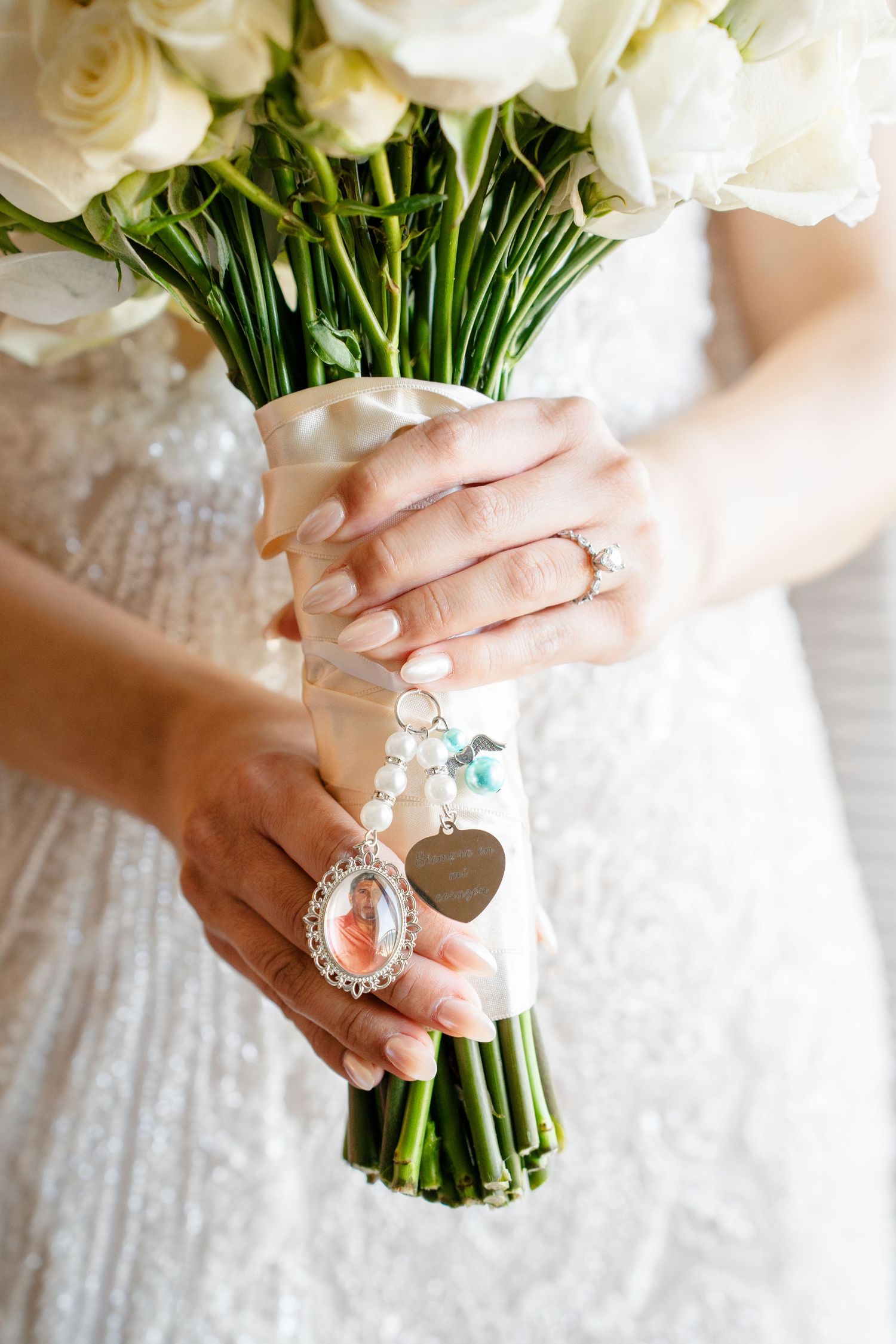 Bride holding white rose bouquet with pearl charm, photo locket, and heart pendant memorial charm, wearing diamond ring.