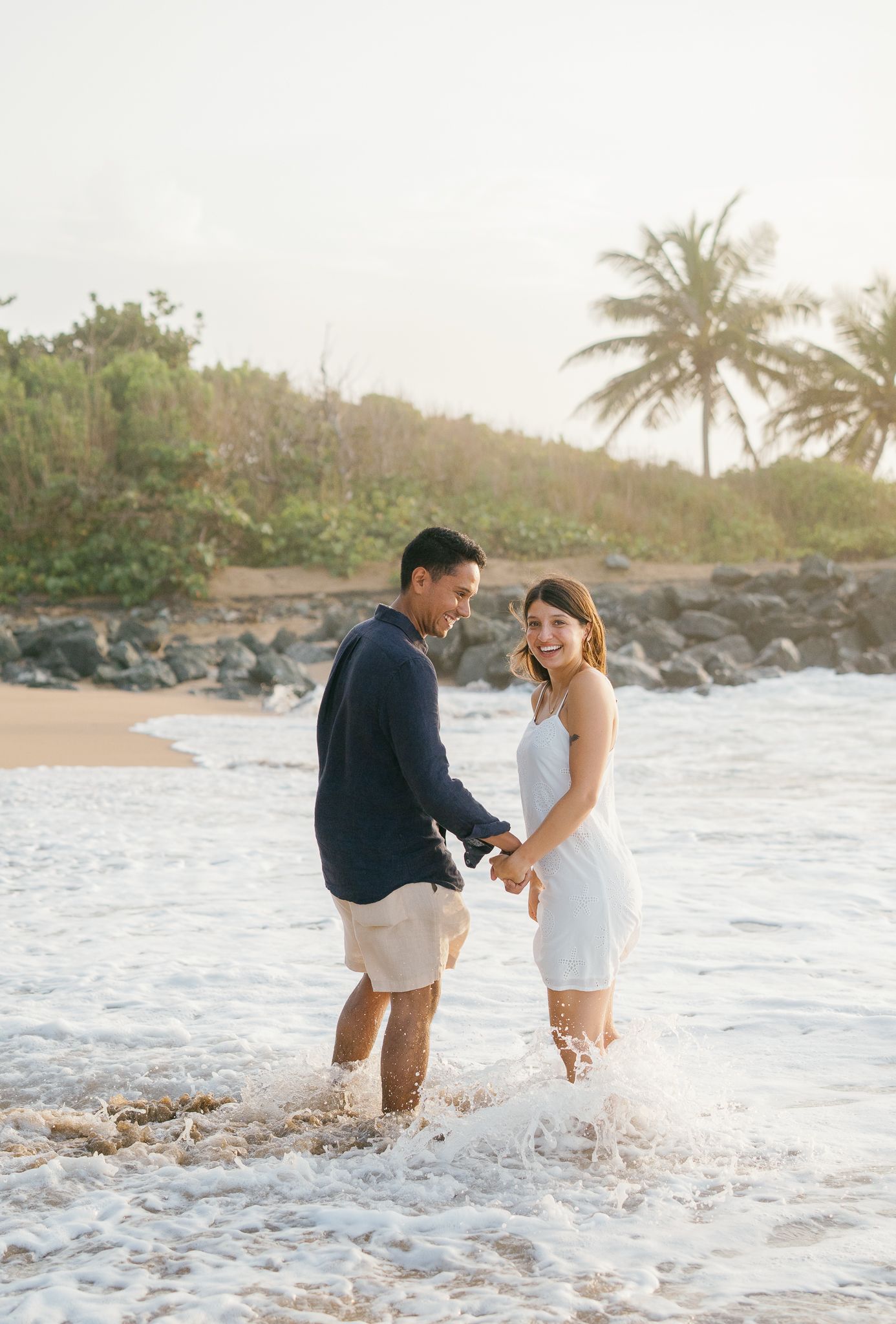 A romantic couple stands together in shallow ocean waves during golden hour at a tropical beach with palm trees.