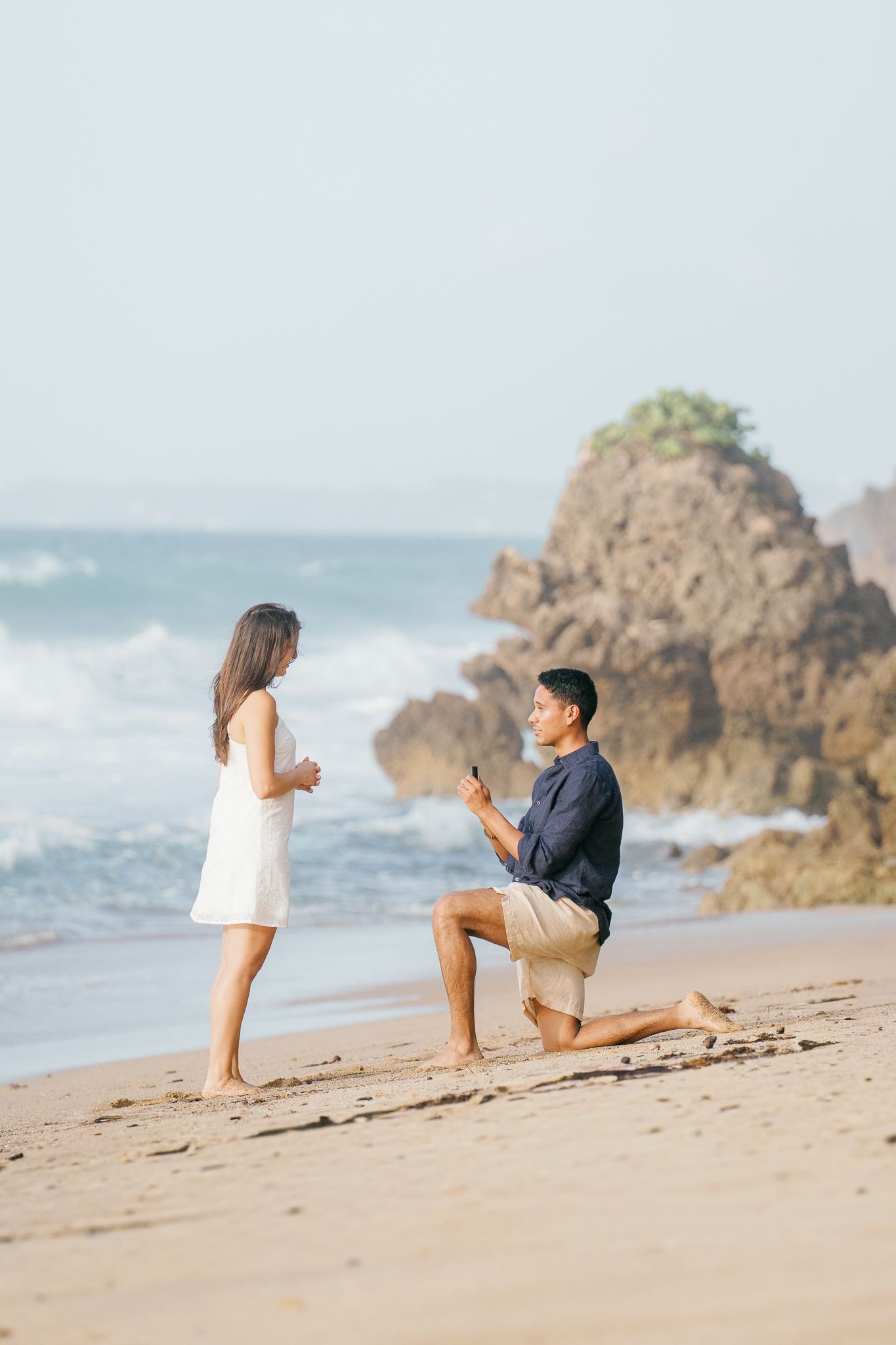 A romantic proposal scene on a scenic beach with crashing waves and rocky cliffs in the background.