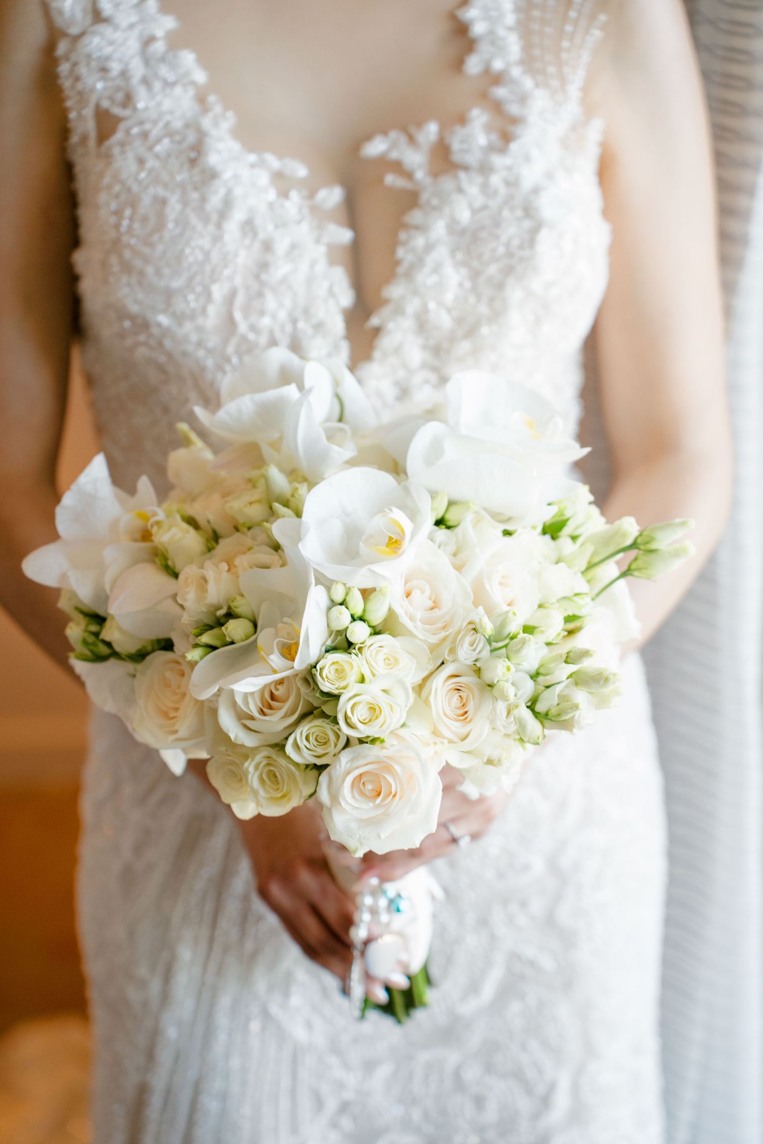 Bride in lace wedding gown holding elegant white bouquet of roses, orchids, and freesia flowers.