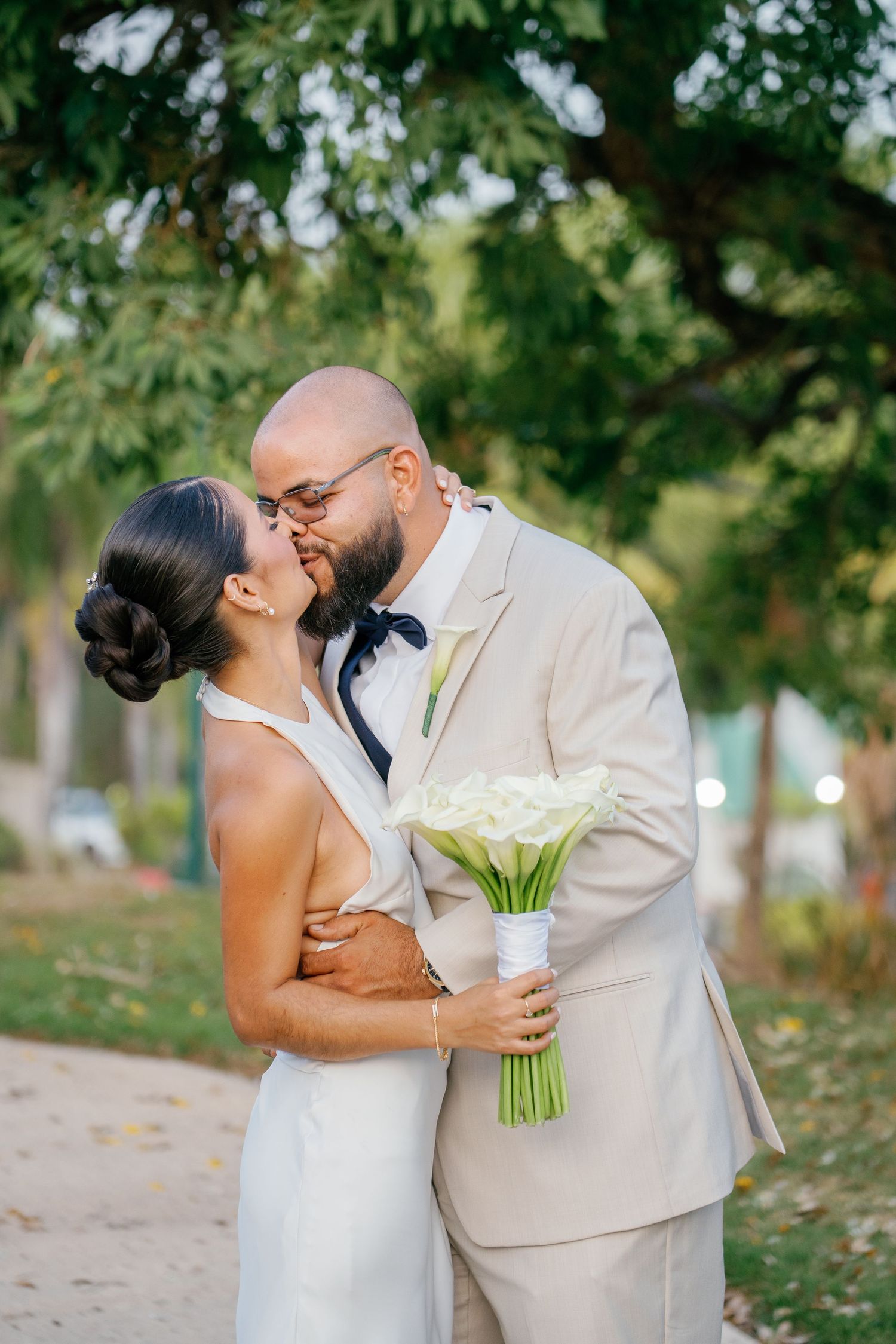 Bride and groom share a romantic kiss outdoors, bride holding white calla lily bouquet, groom in beige suit with navy bow tie.