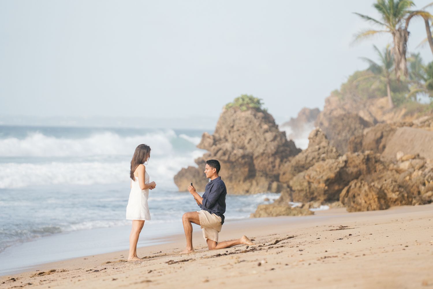 A romantic beach proposal scene with waves crashing against rocky cliffs and palm trees in the background.