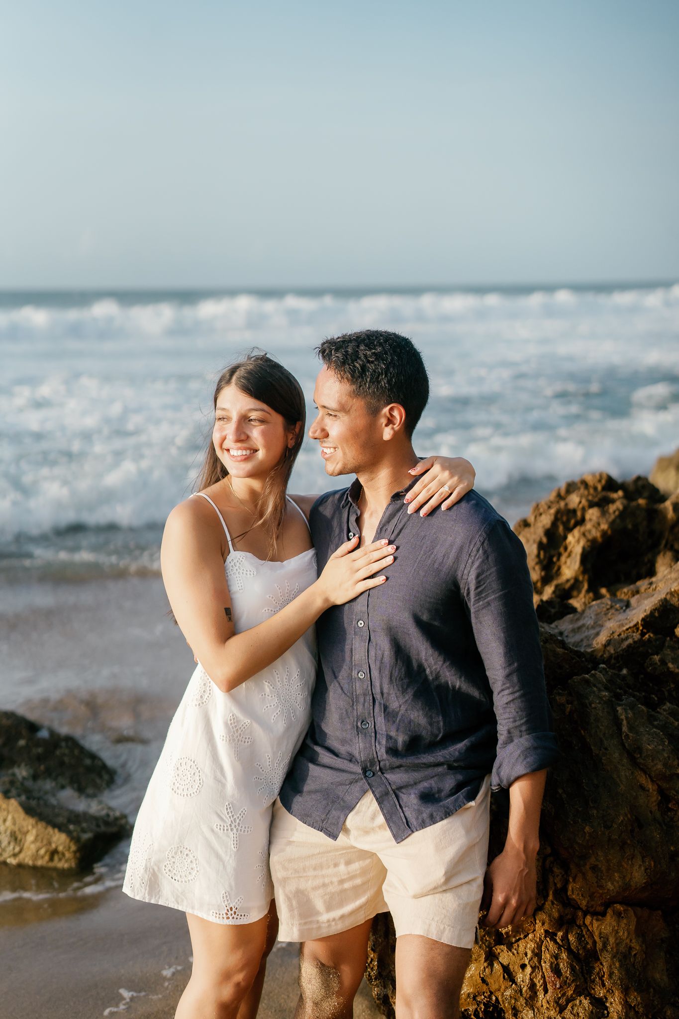 A couple shares an intimate moment on a rocky beach with crashing waves in the background.