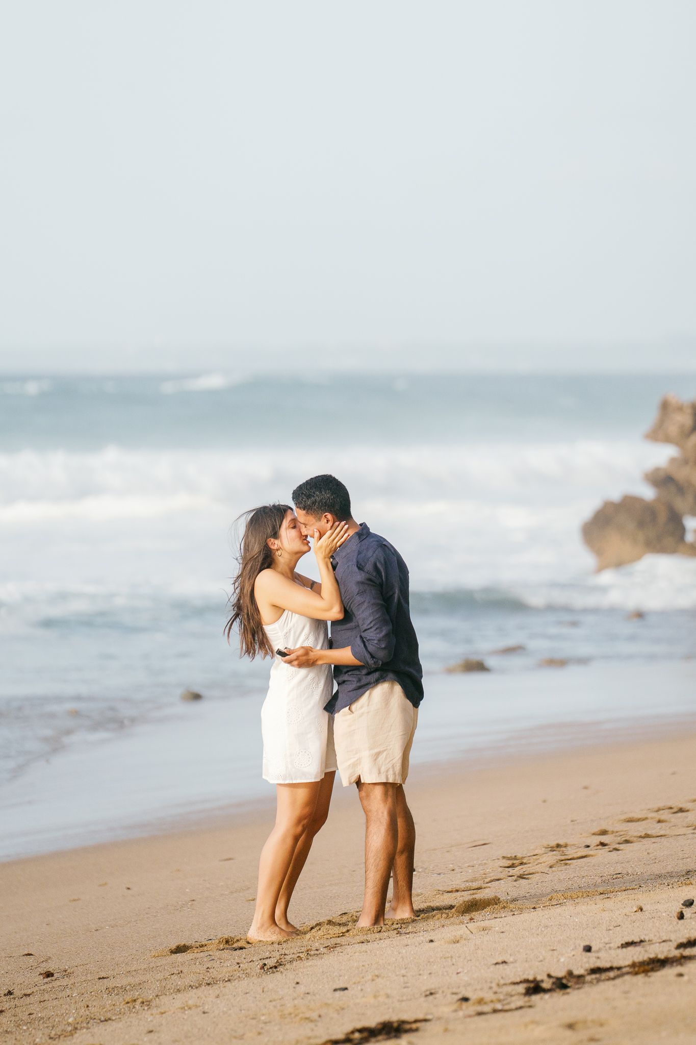 A romantic couple embraces and kisses on a sandy beach with ocean waves and rocks in the background.