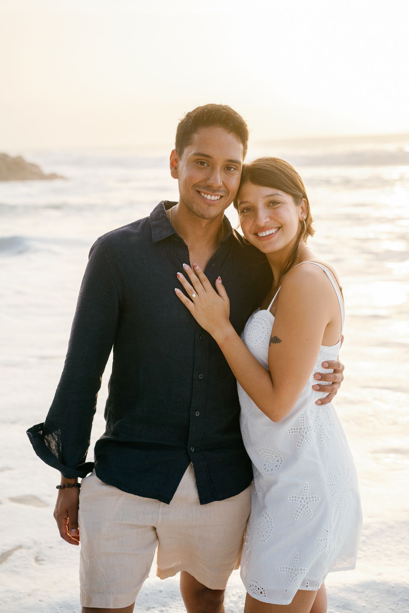 A couple poses together on a sunny beach in coordinated black and white outfits during a romantic photoshoot.