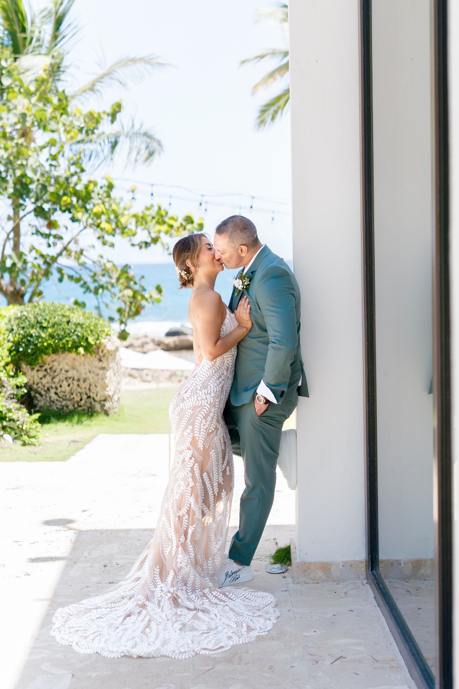 Elegant wedding couple embracing against white wall with ocean view and palm trees.