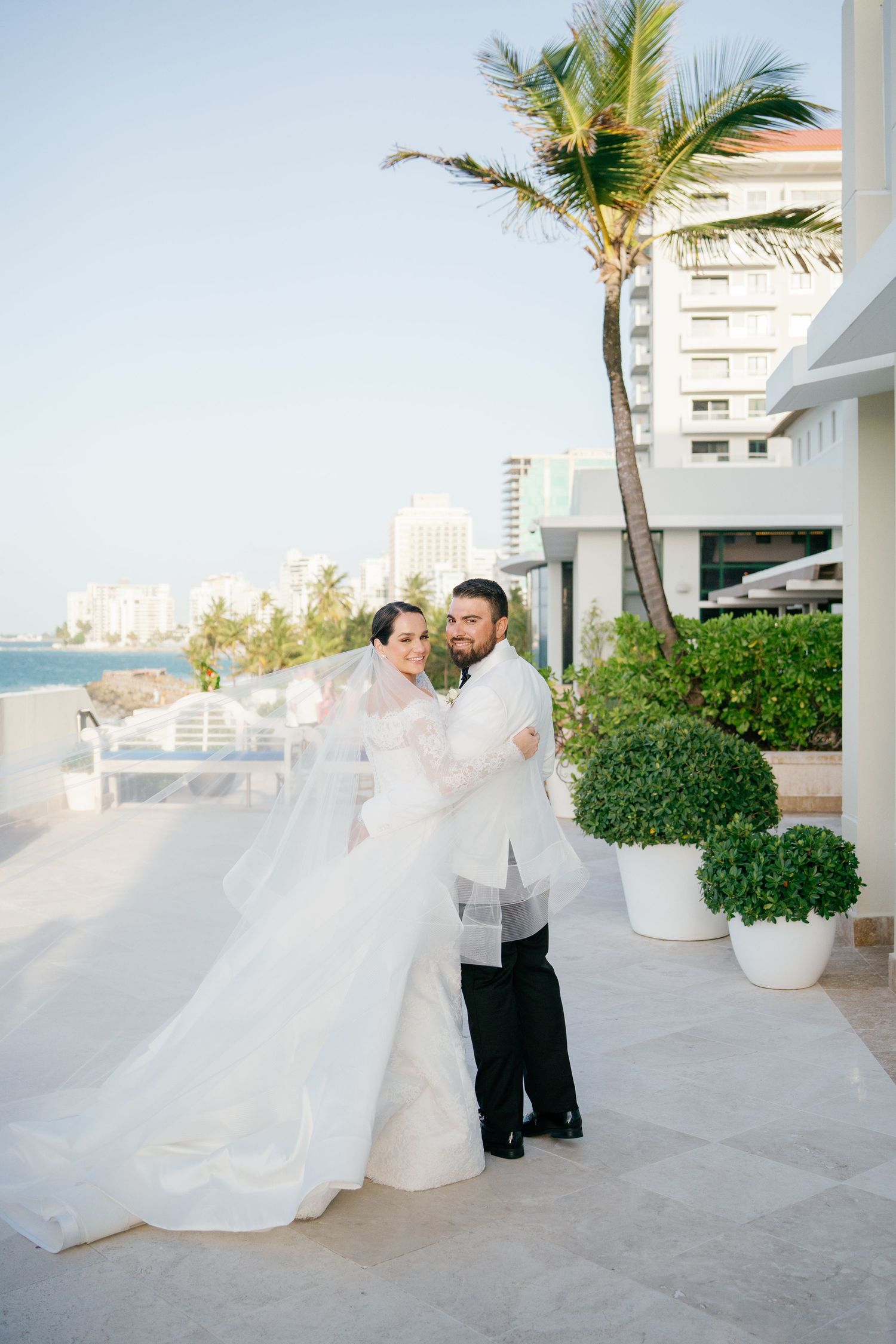 Bride and groom pose on a waterfront terrace with flowing veil, palm trees, and city skyline backdrop on their wedding day.