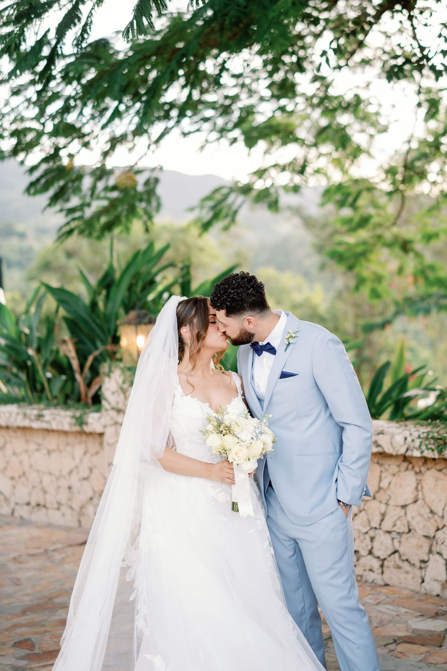 A bride and groom share a romantic forehead kiss outdoors, she in a white gown with veil, he in a light blue suit.